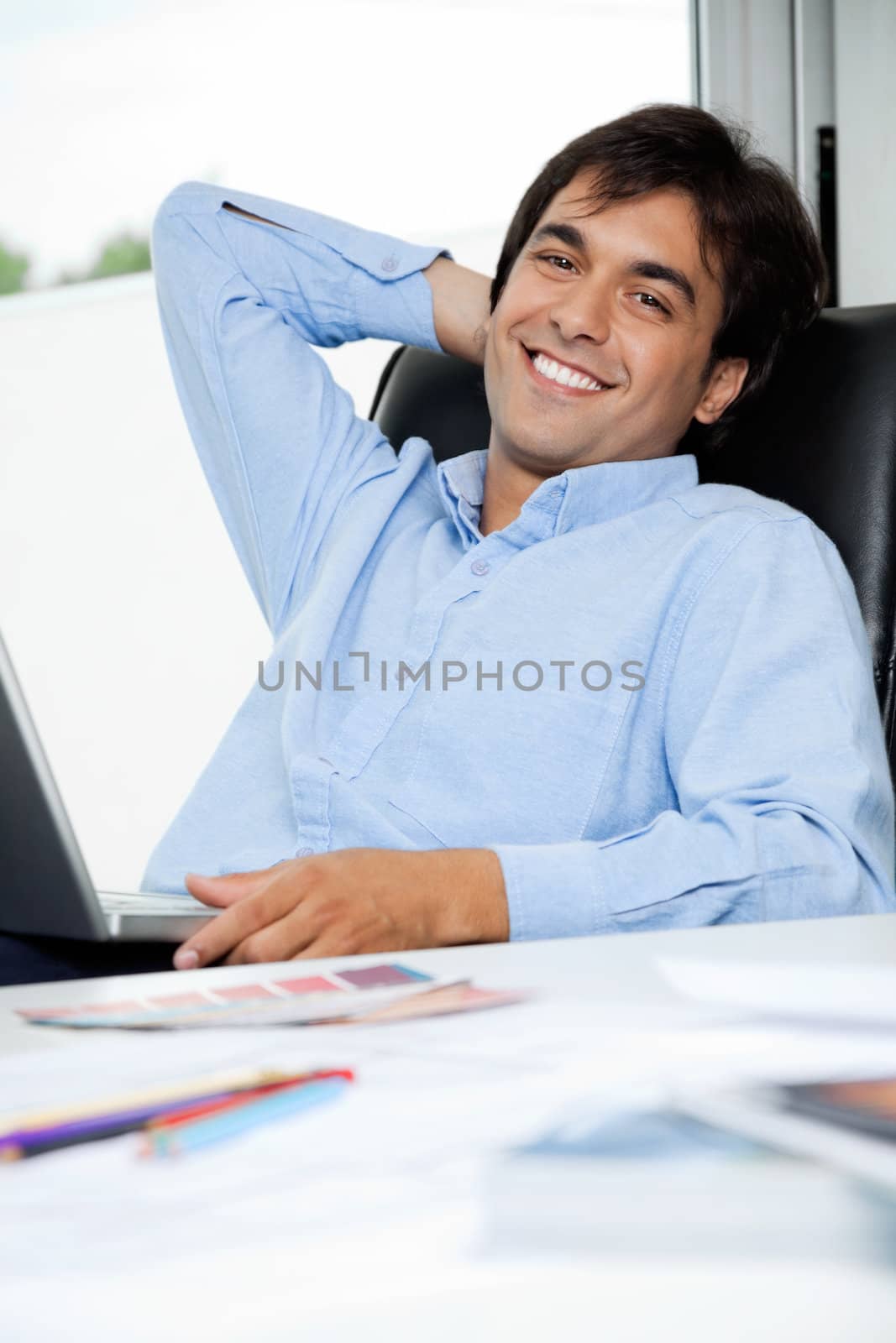 Portrait of relaxed young male interior designer with laptop sitting in office chair