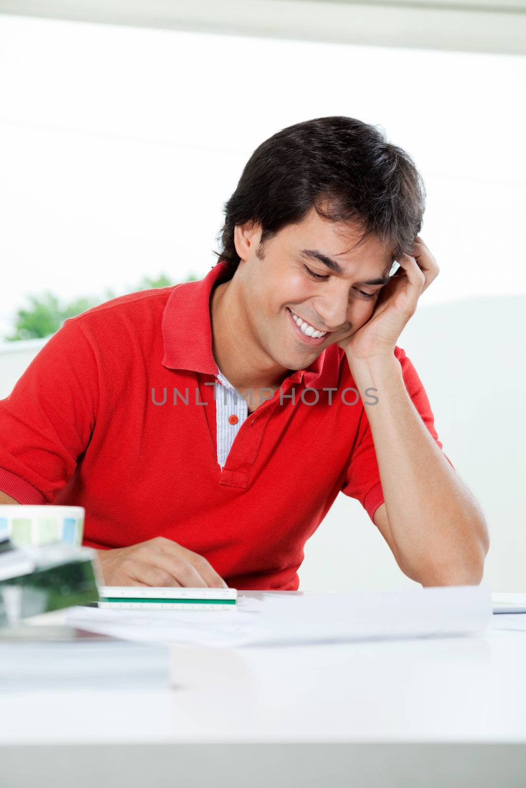 Happy young male architect in casual red t-shirt looking down while sitting at table