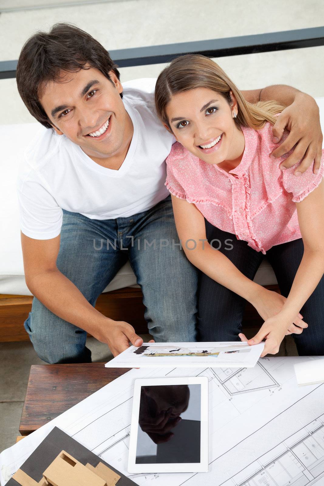 Portrait of young couple holding house plans with tablet PC, blueprint and model structure on table