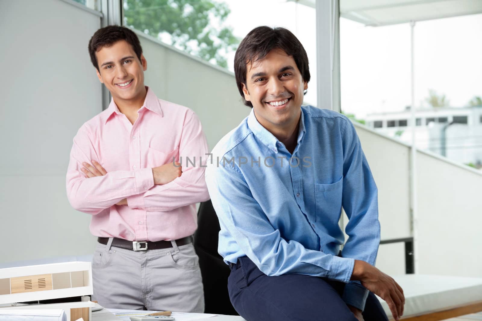 Portrait of young male architect smiling while colleague stands with arms crossed in background