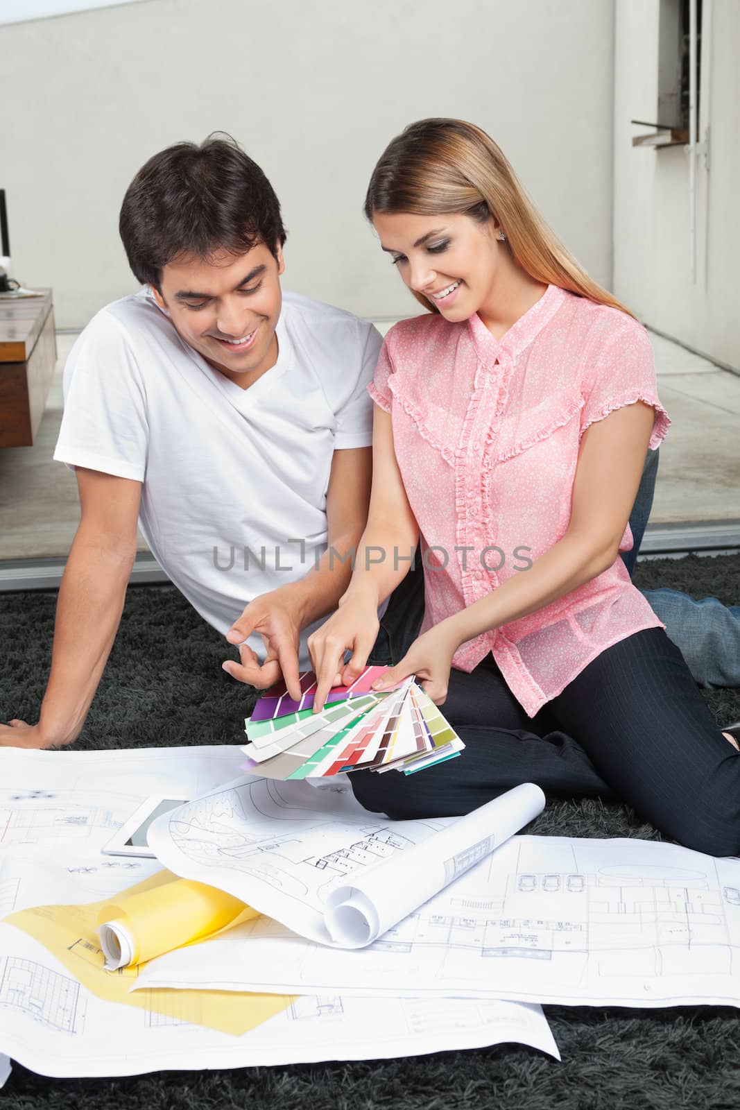 Young couple choosing colors from swatches while sitting on rug with blueprints