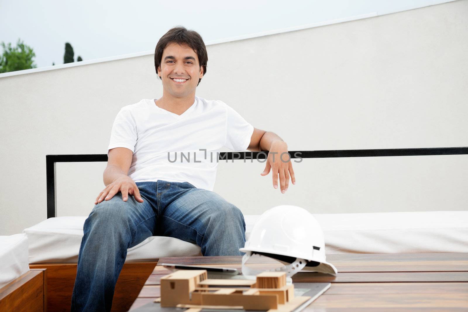 Portrait of a relaxed young male architect with hardhat and model structure on table