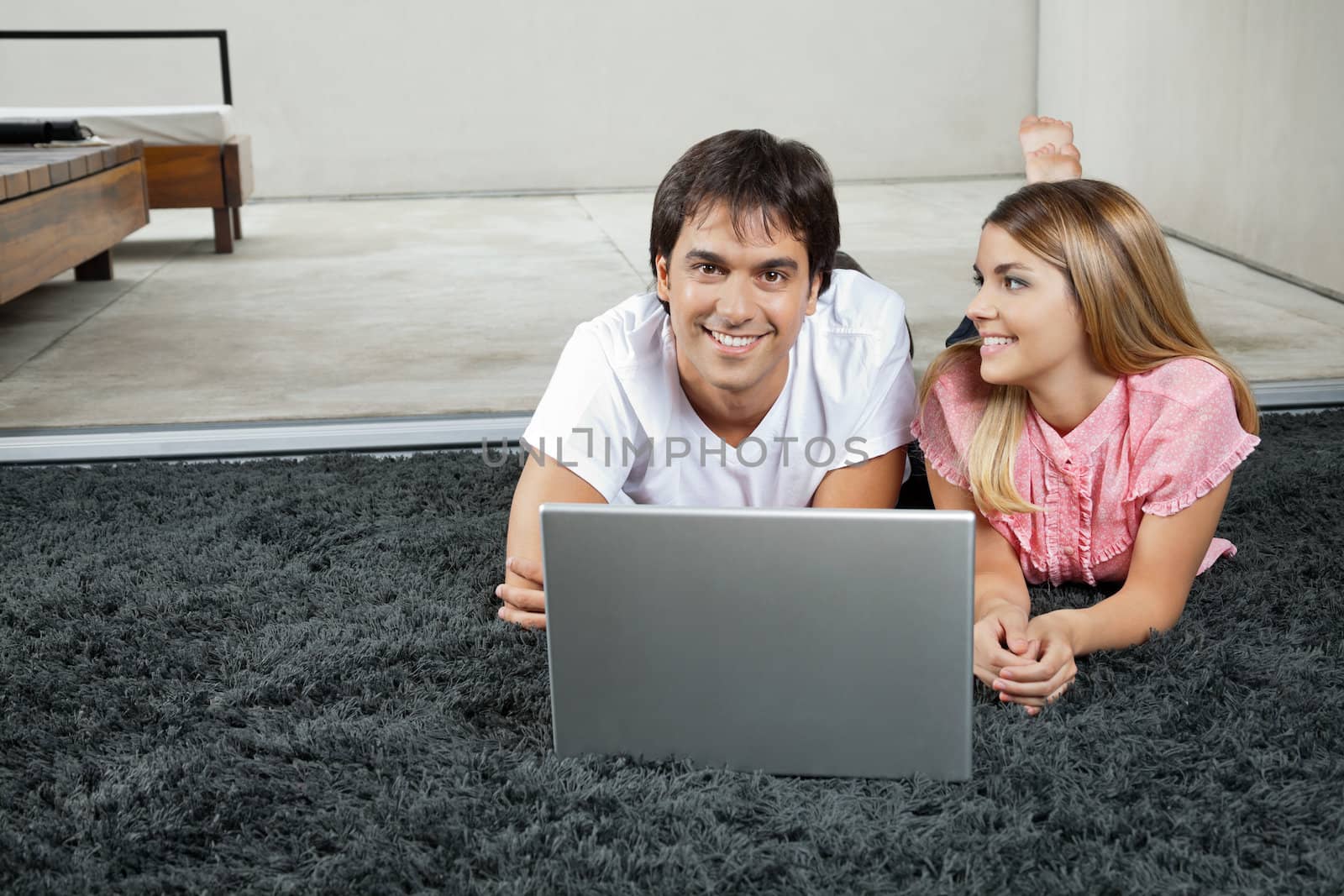 Portrait of happy young man lying on rug with laptop while woman looking at him