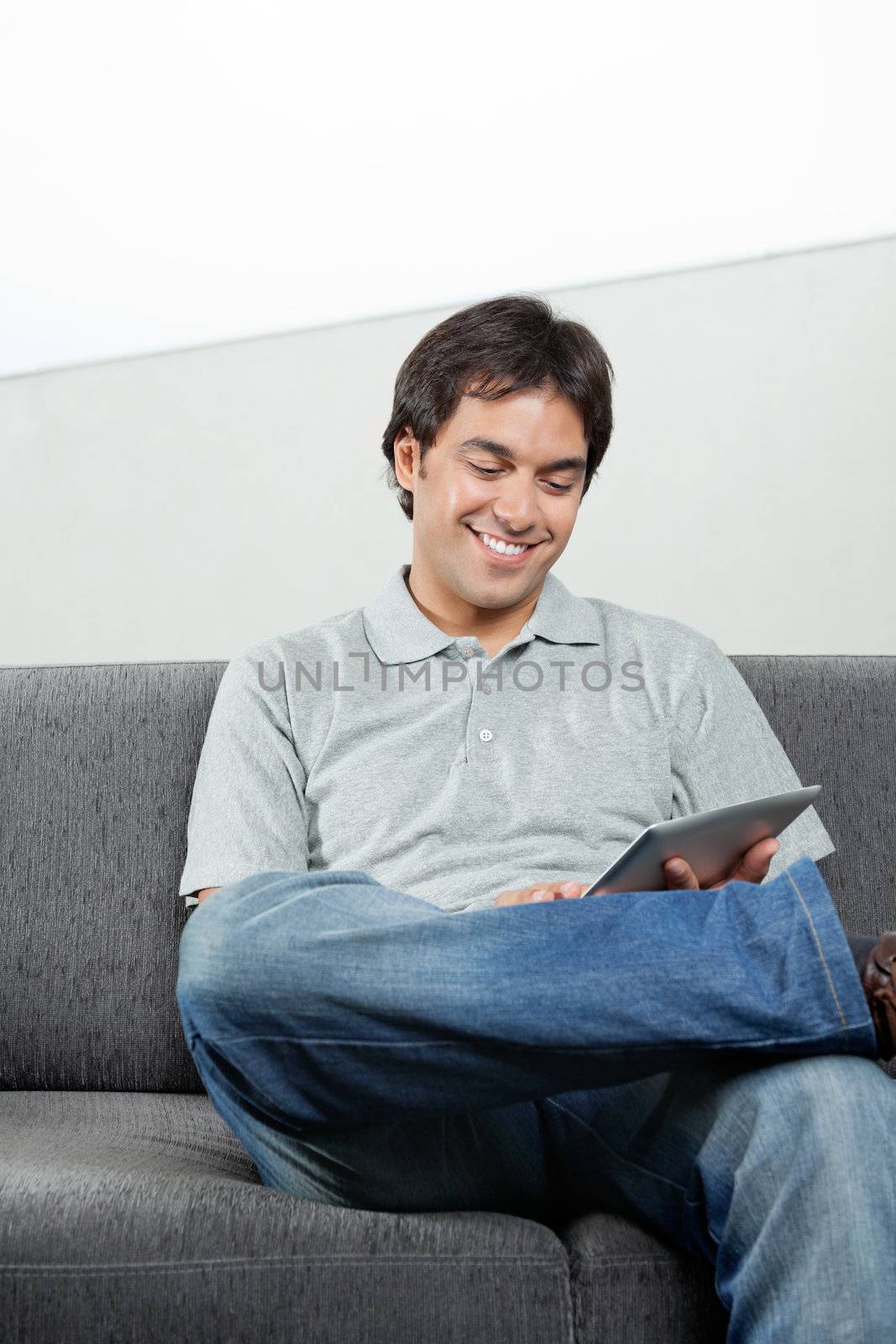 Happy young male in casual wear browsing internet on digital tablet while sitting on sofa