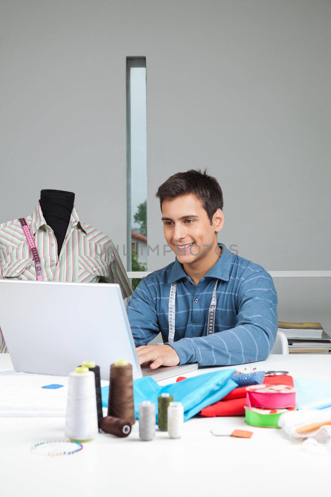 Happy male tailor using laptop while sitting by table with dressmaking accessories