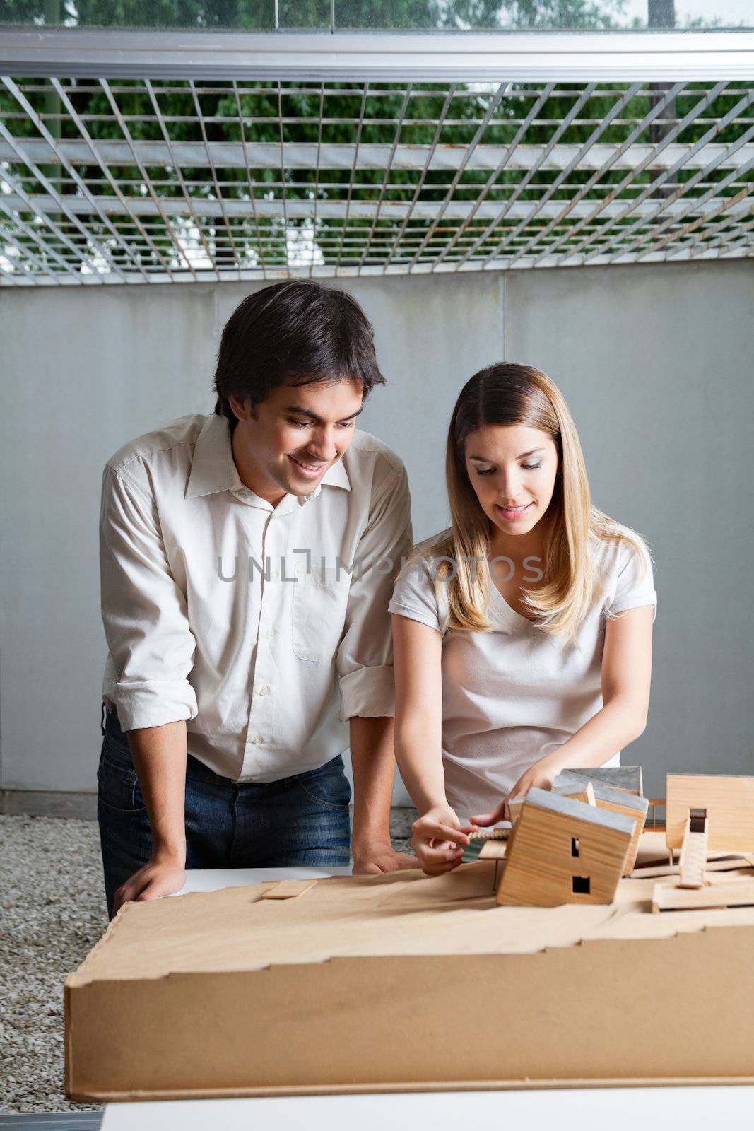 Young male architect standing beside colleague working on model house