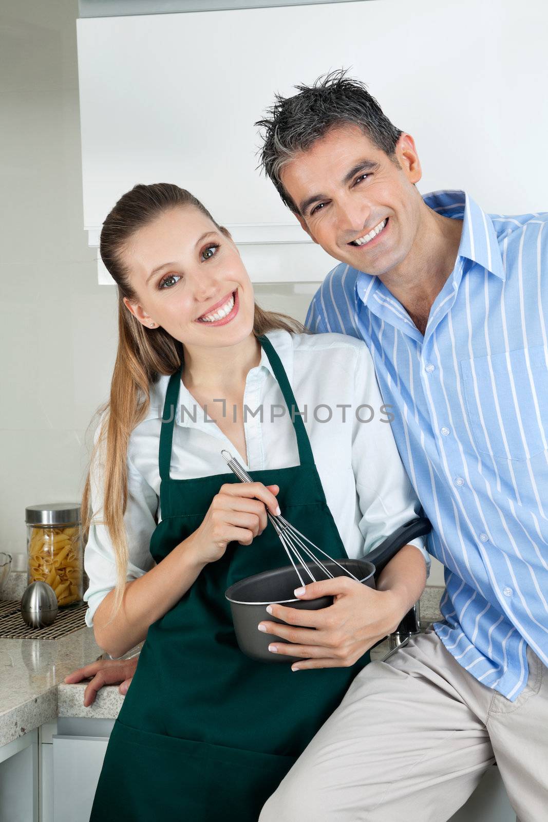 Portrait of happy young woman holding pan and whisk while standing with boyfriend in kitchen