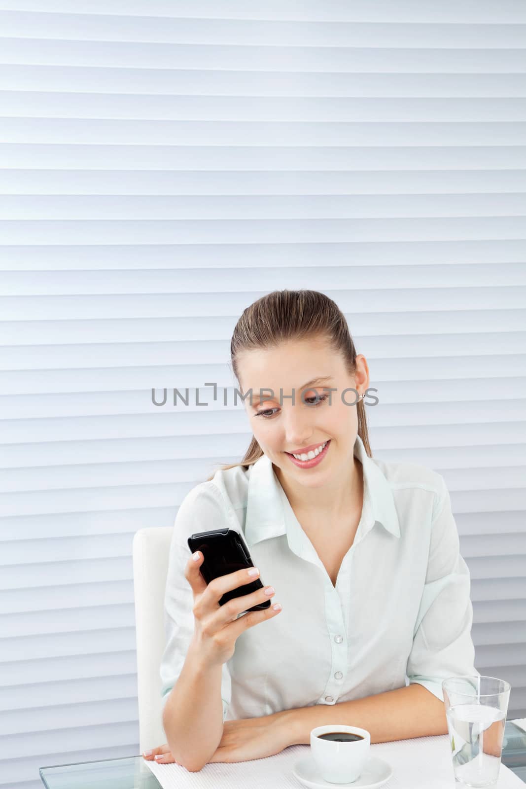 Beautiful young businesswoman reading text message on cell phone while sitting at table with tea and glass of water