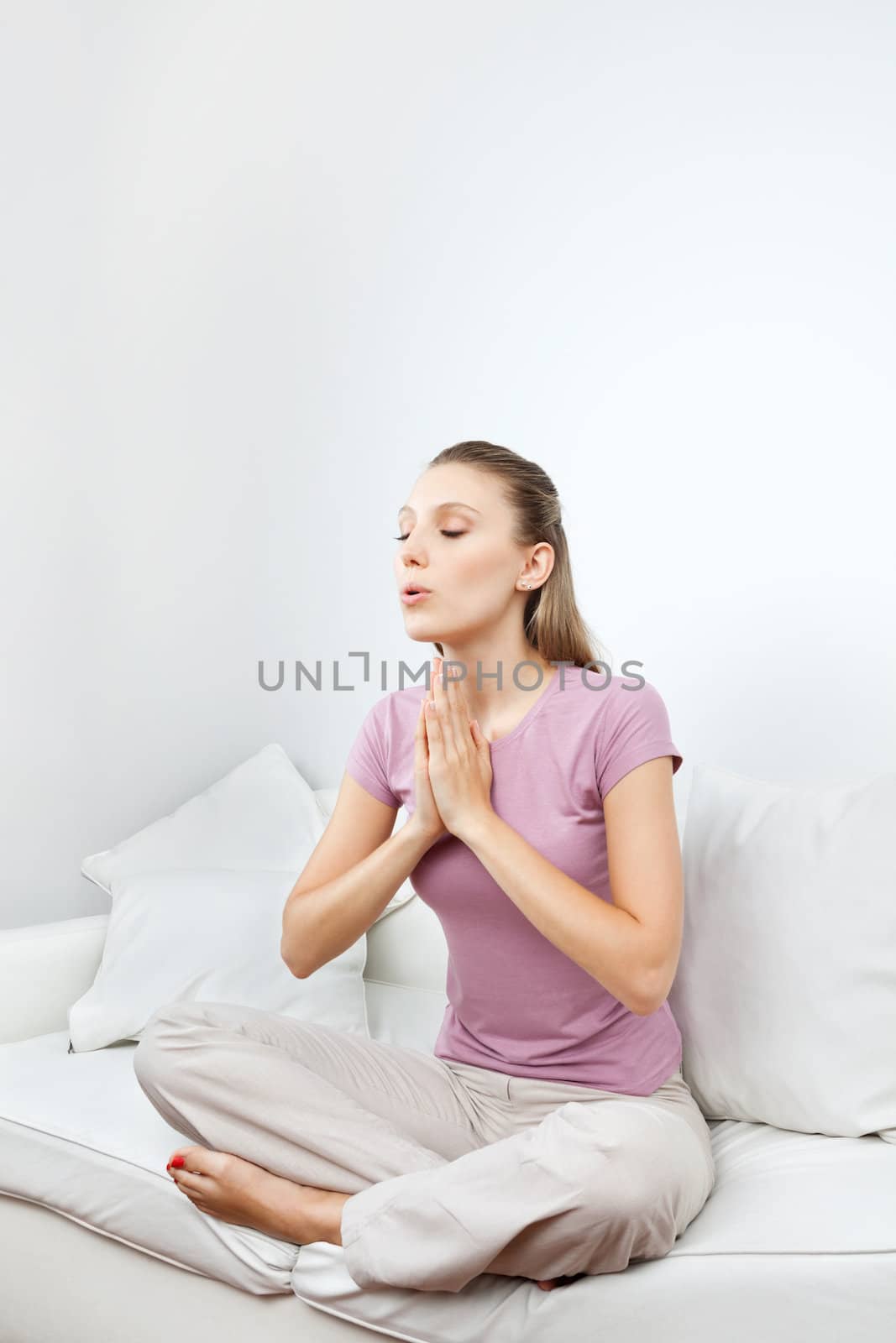 Young woman in casual wear meditating with hands clasped on sofa at home