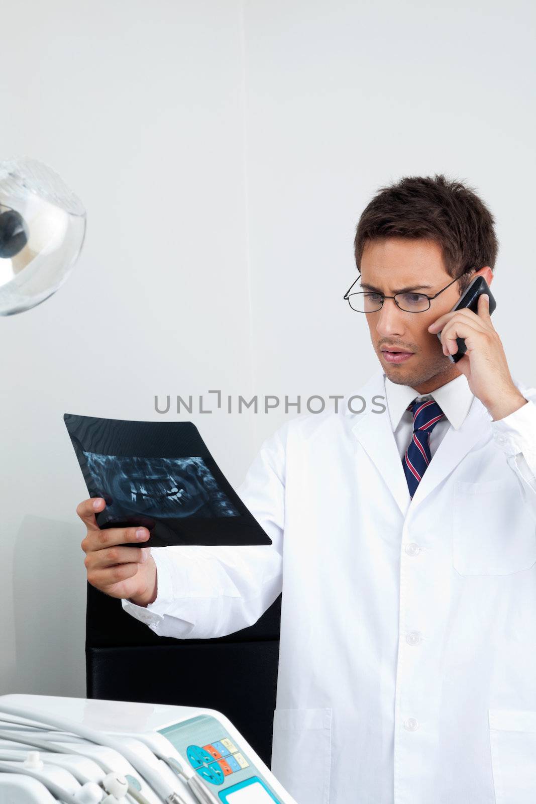 Young male doctor looking at X-ray report while using mobile phone in dental clinic