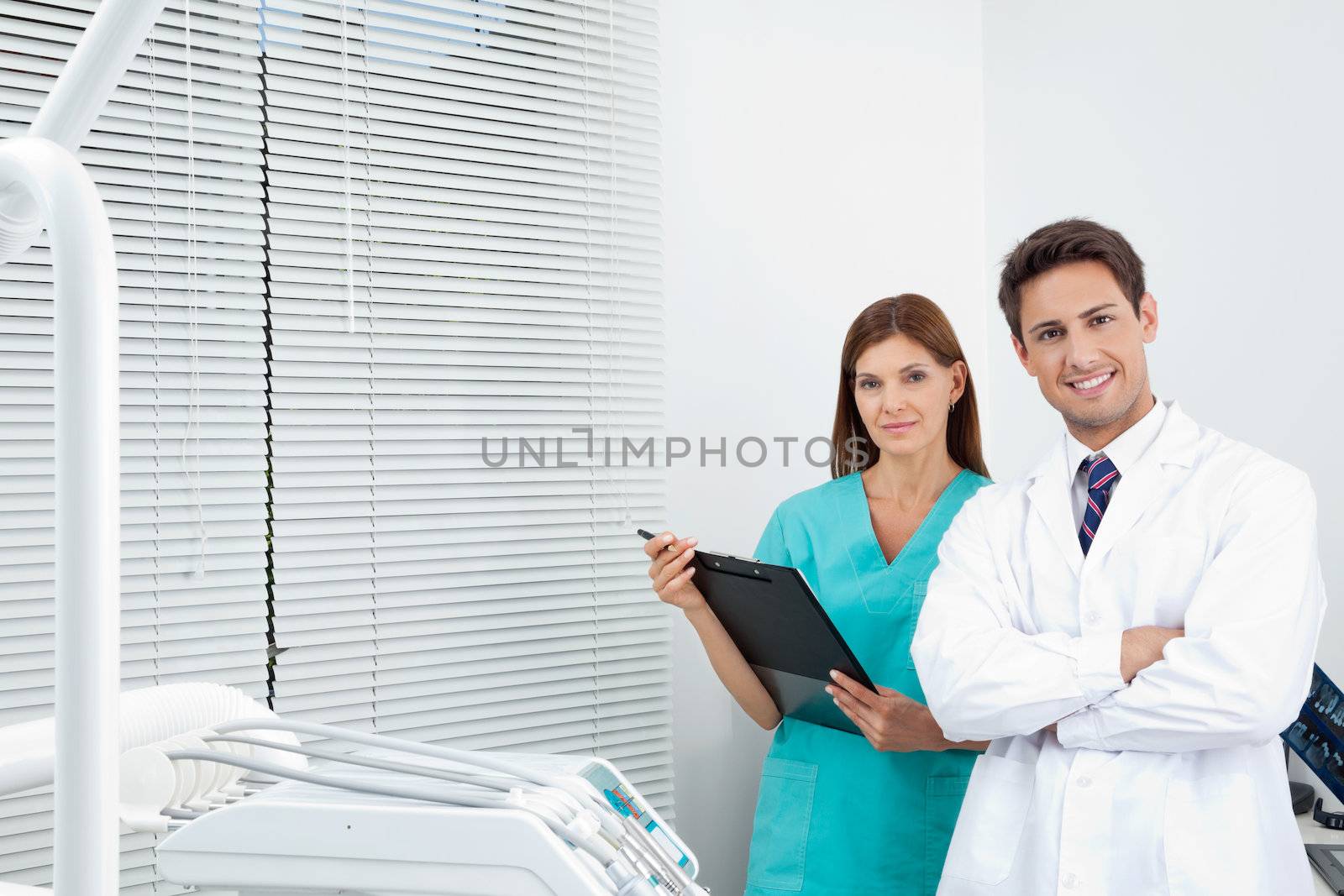 Portrait of happy young doctor and female assistant with clipboard in dental clinic