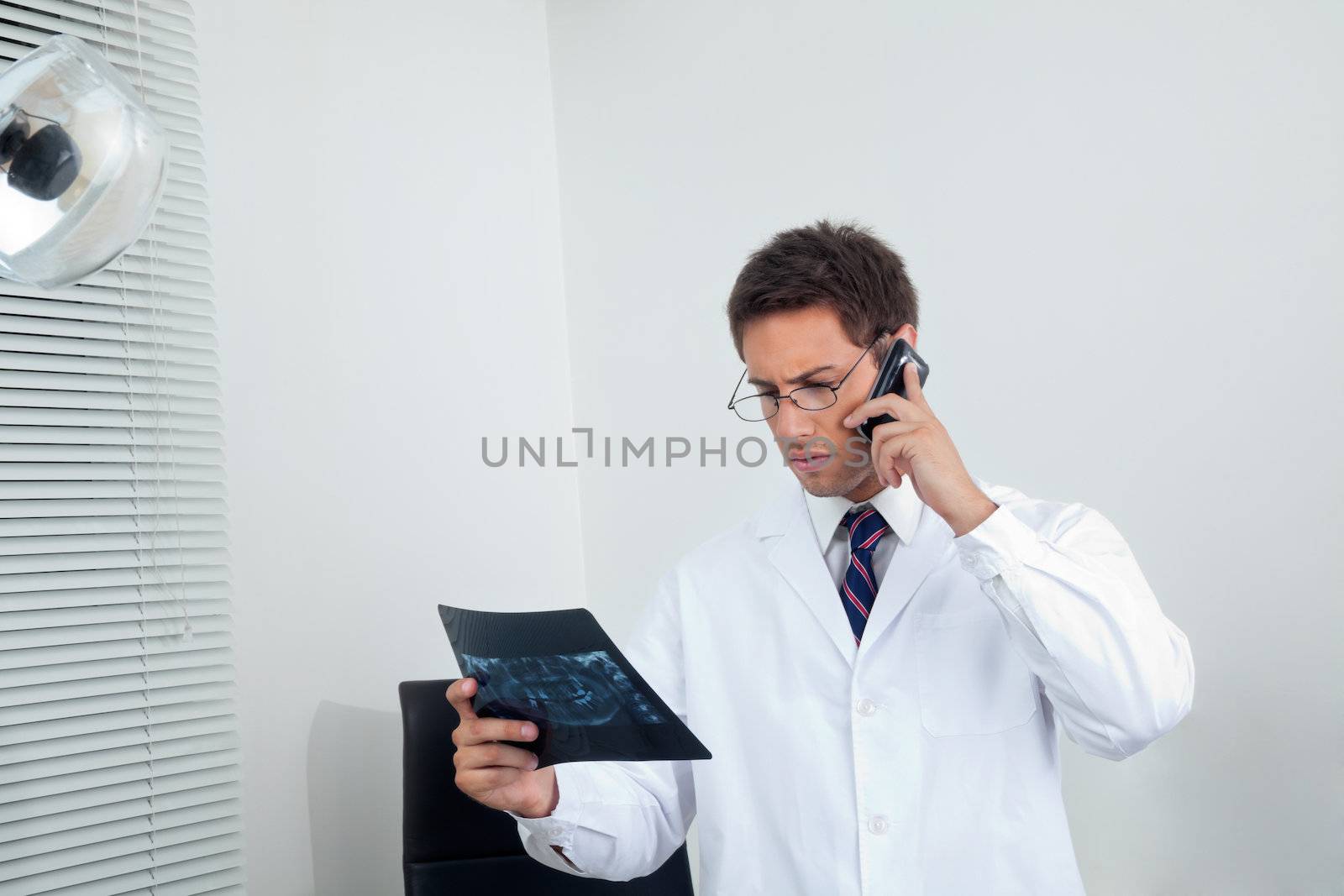 Young male dentist looking at X-ray report while using cellphone in clinic