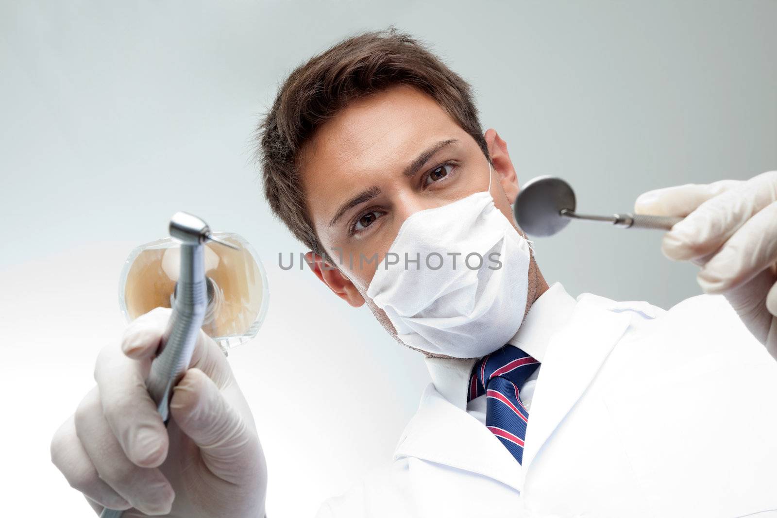 Portrait of young male dentist wearing surgical mask while holding angled mirror and drill