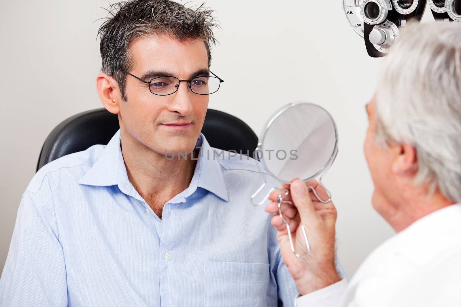 Man in stylish reading glasses while looking at mirror with optometrist in the clinic