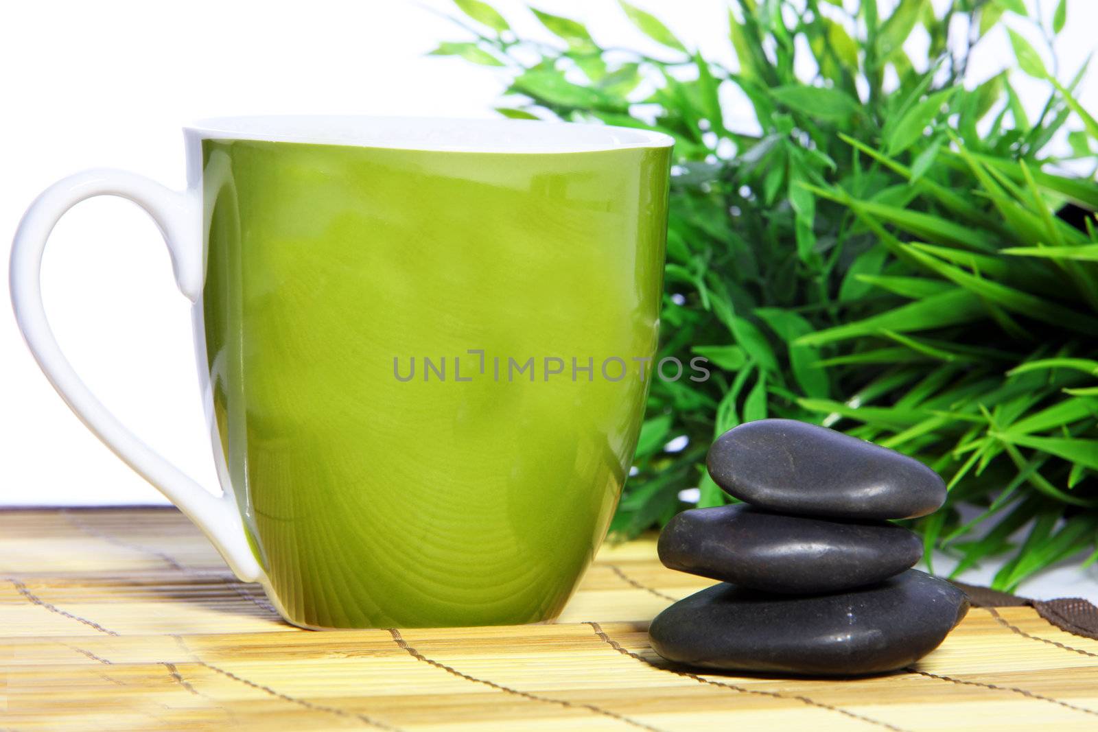 Pretty green glazed ceramic mug and spa massage stones standing on a reed mat in front of fresh green bamboo leaves in a spa, health and wellbeing concept