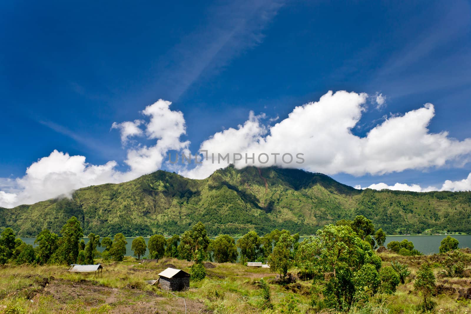 Bali landscape wit two small rural huts in a lush green valley with a mountain backkdrop under a cloudy blue sky and sunshine