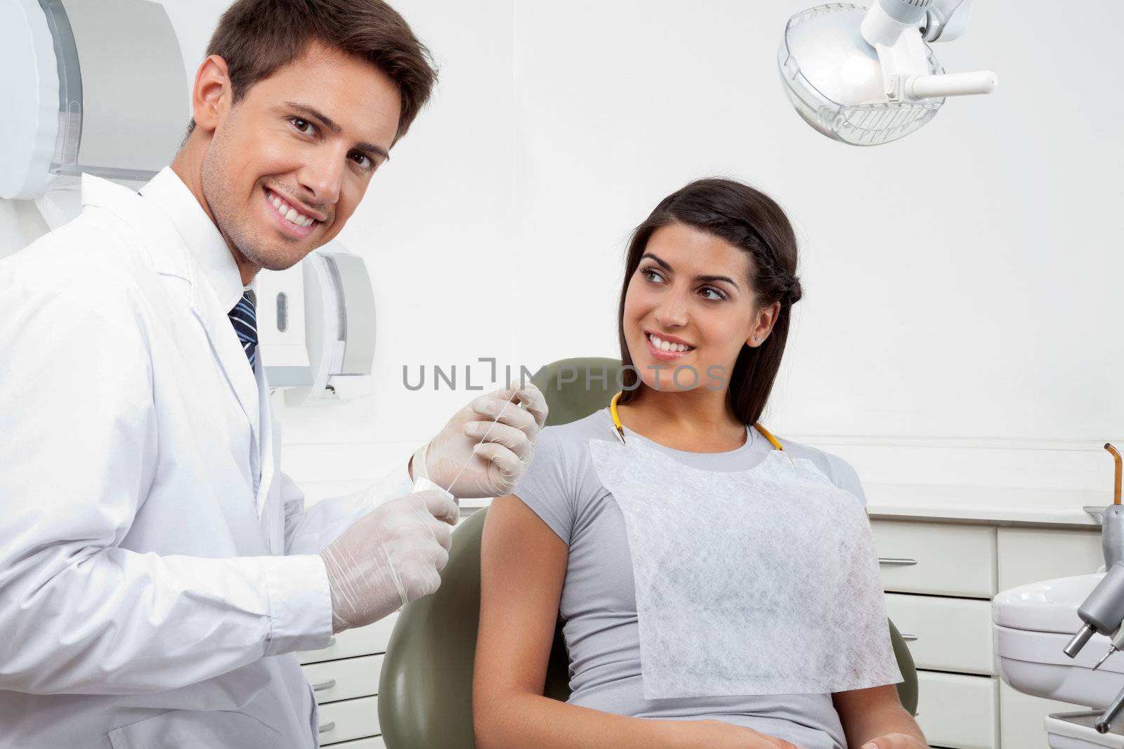 Portrait of happy young male dentist holding thread while patient looking at him in clinic