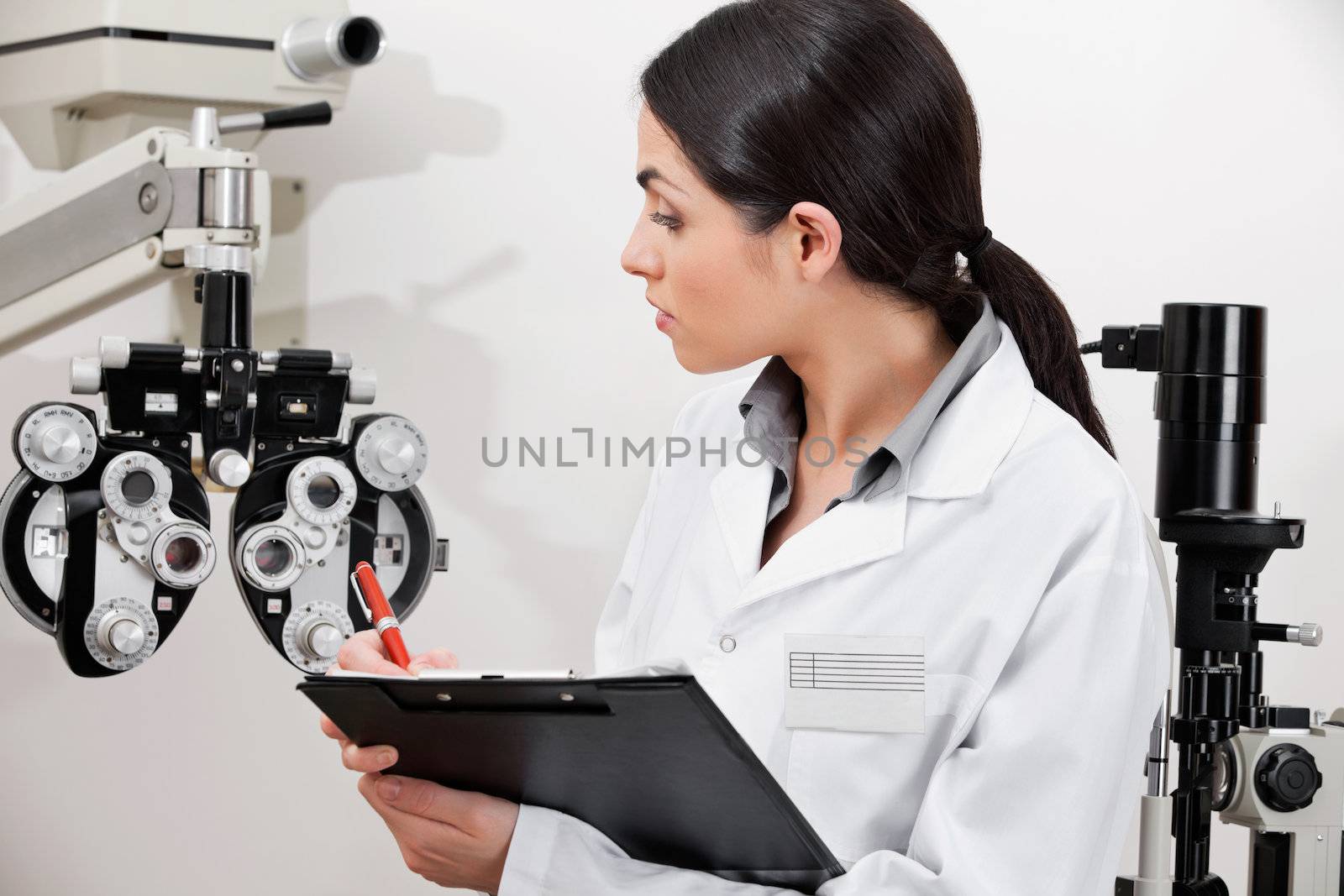 Female eye specialist noting down on clipboard while looking at phoropter