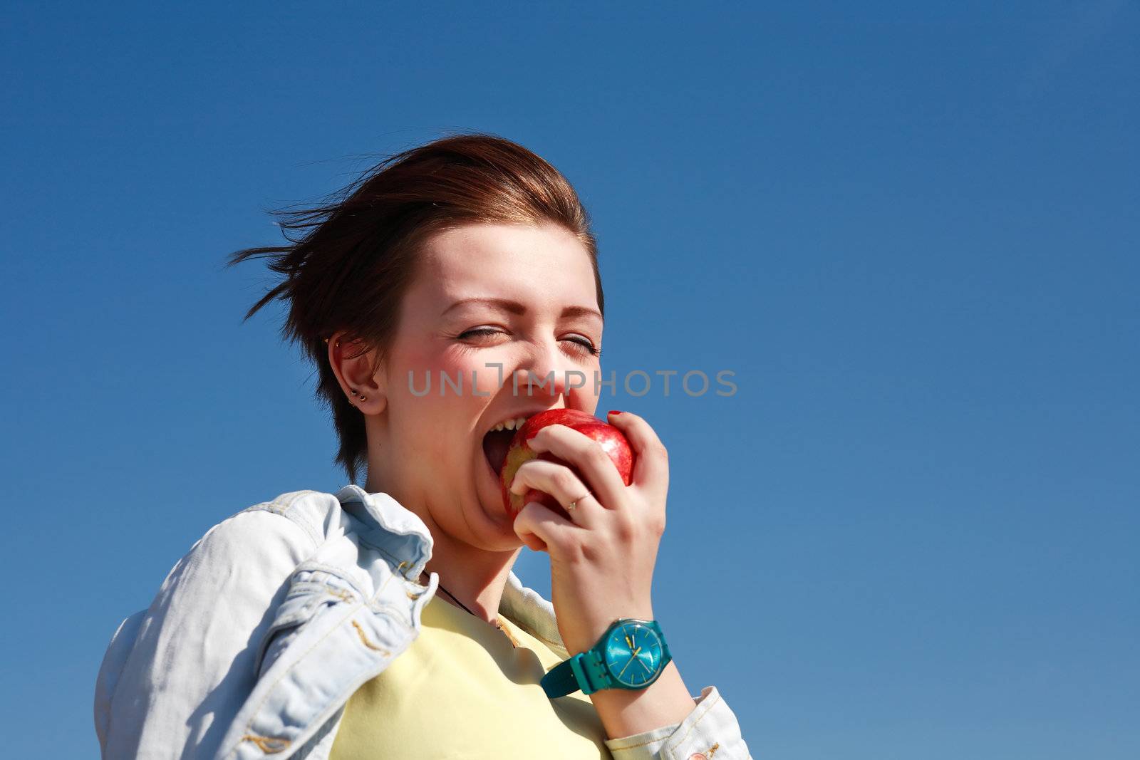 Beautiful young girl eating rich red apple against blue sky background