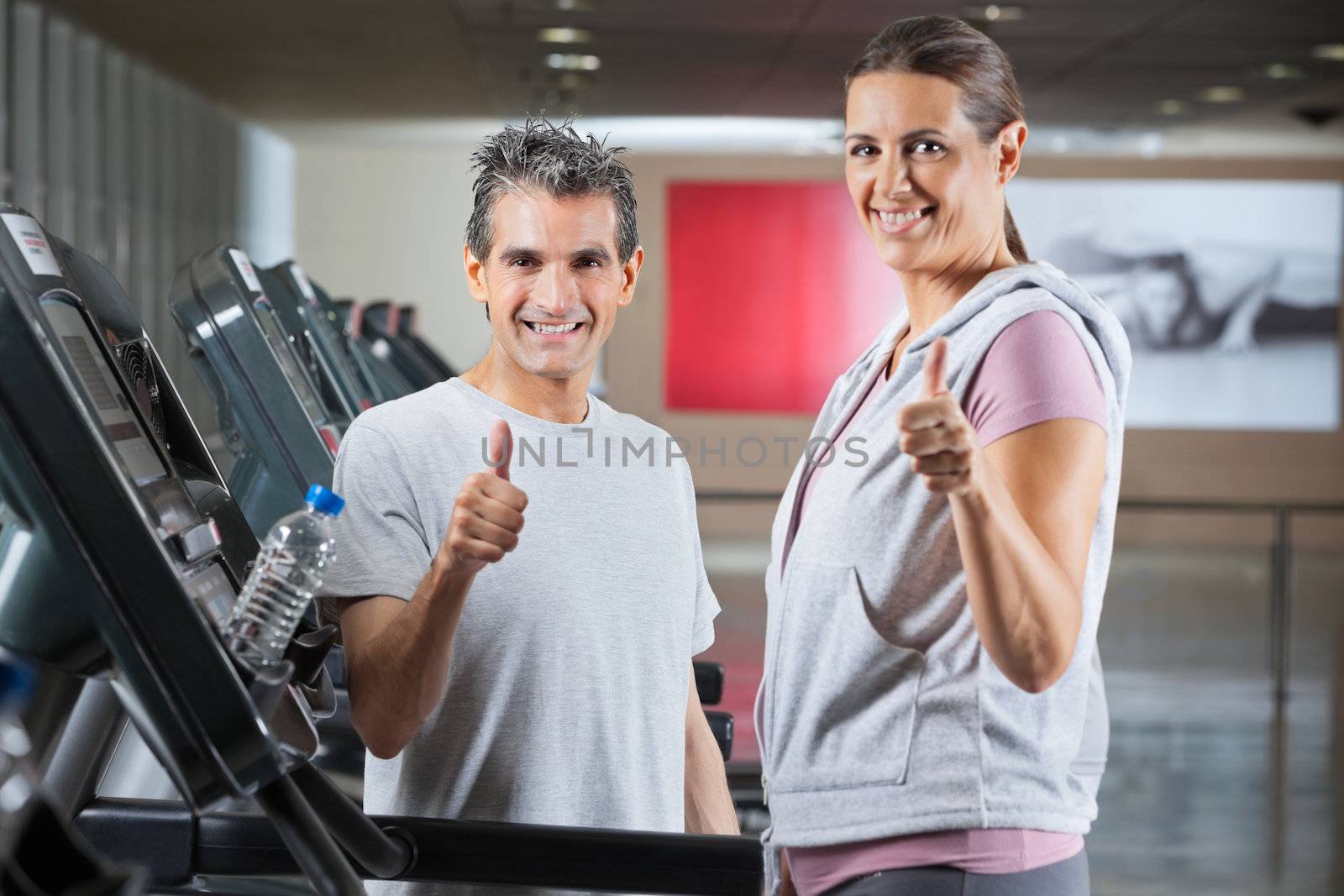 Happy mature instructor and female client showing thumbs up sign in health club - shallow depth of field, focus on trainer