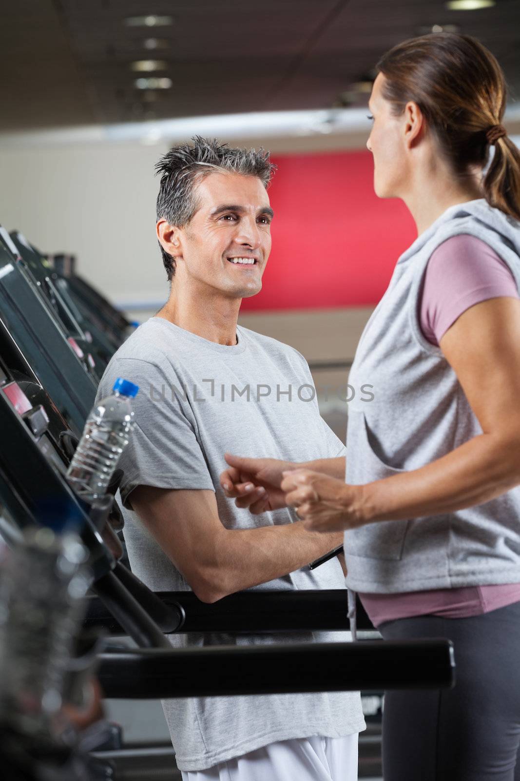 Happy mature instructor looking at female client exercising on treadmill in health center