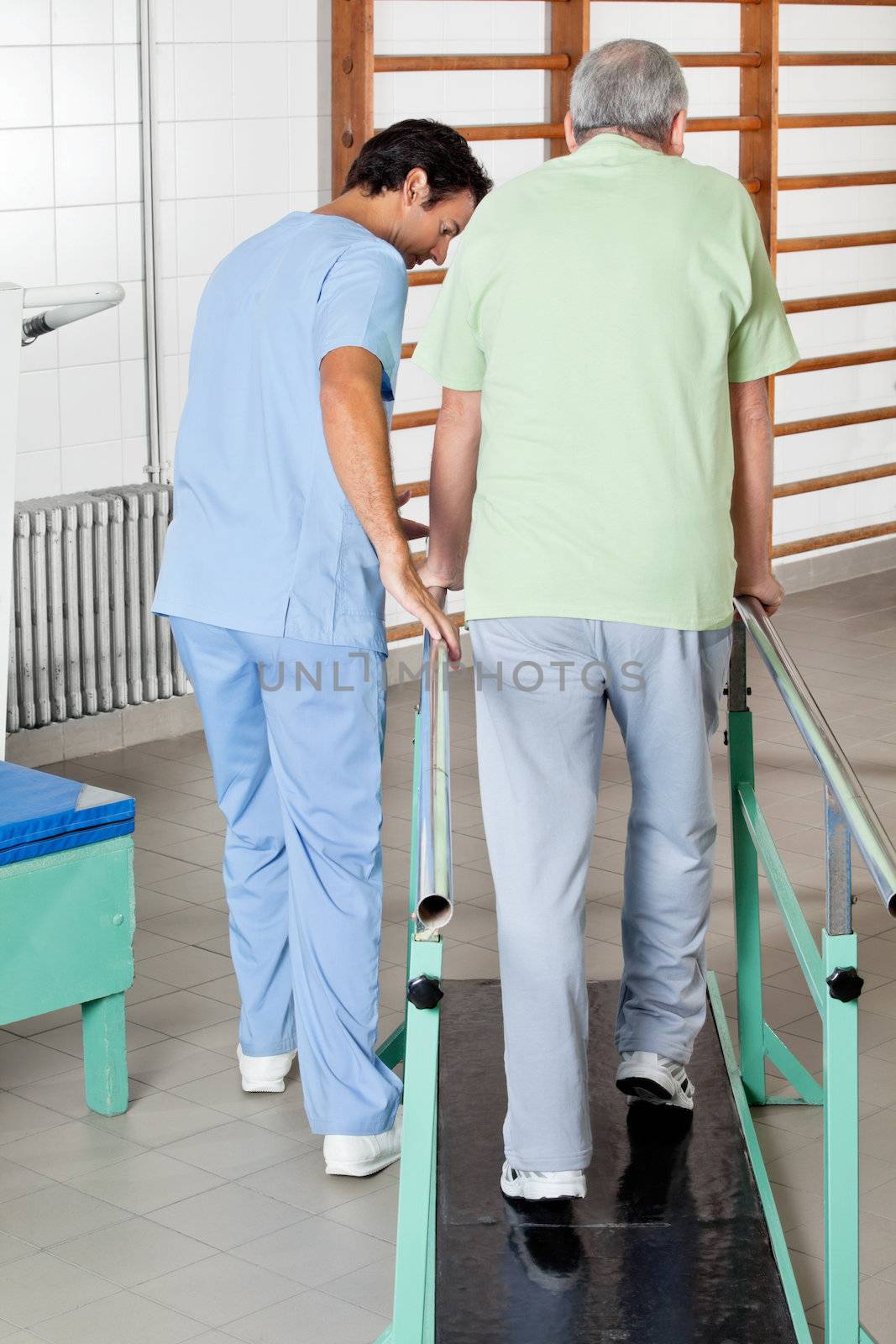 Rear view of male physical therapist assisting senior man to walk with the support of bars at hospital gym