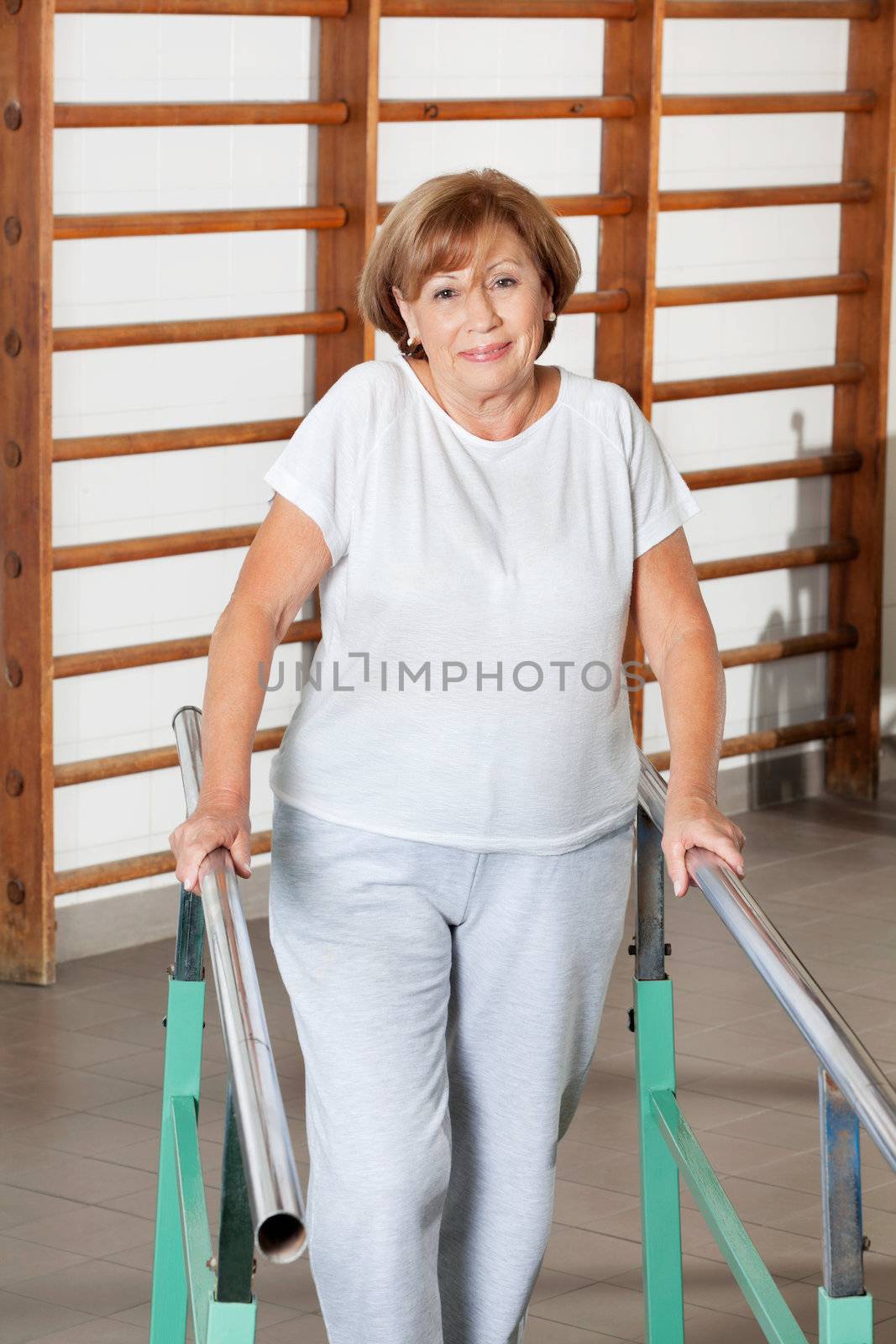 Portrait of a happy senior woman walking with the help of support bars at hospital gym
