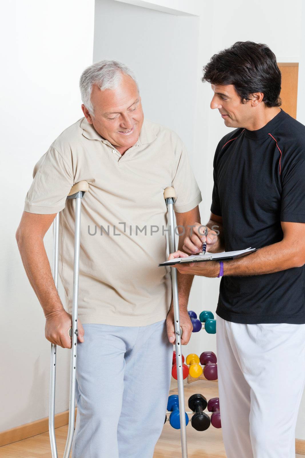 Happy therapist writing report while standing with senior man on crutches
