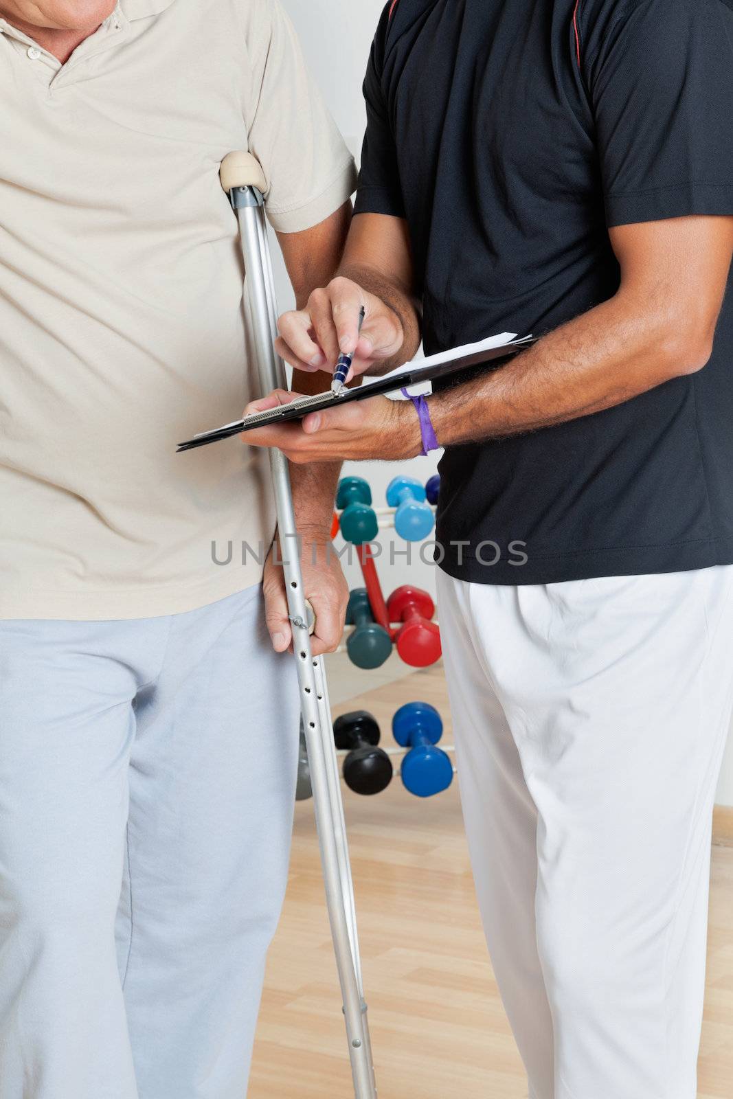 Mid section of trainer holding clipboard while standing with senior man on crutches