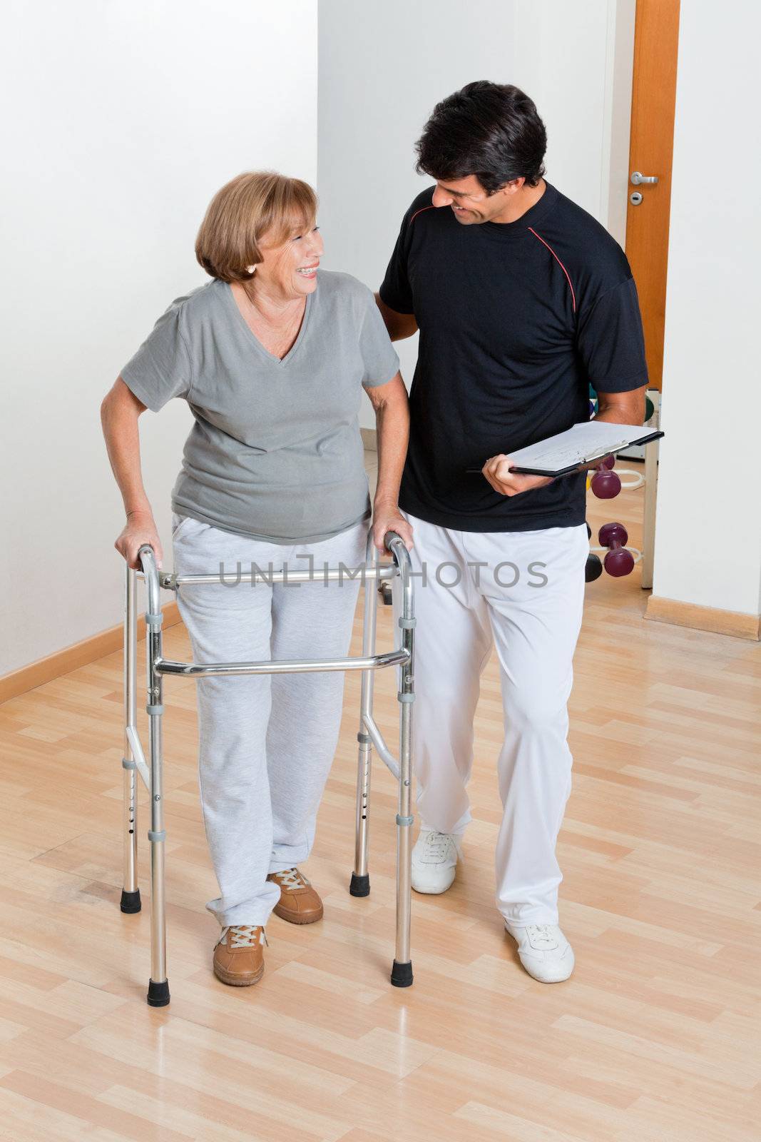 Full length of a trainer holding clipboard and looking at senior woman using walker
