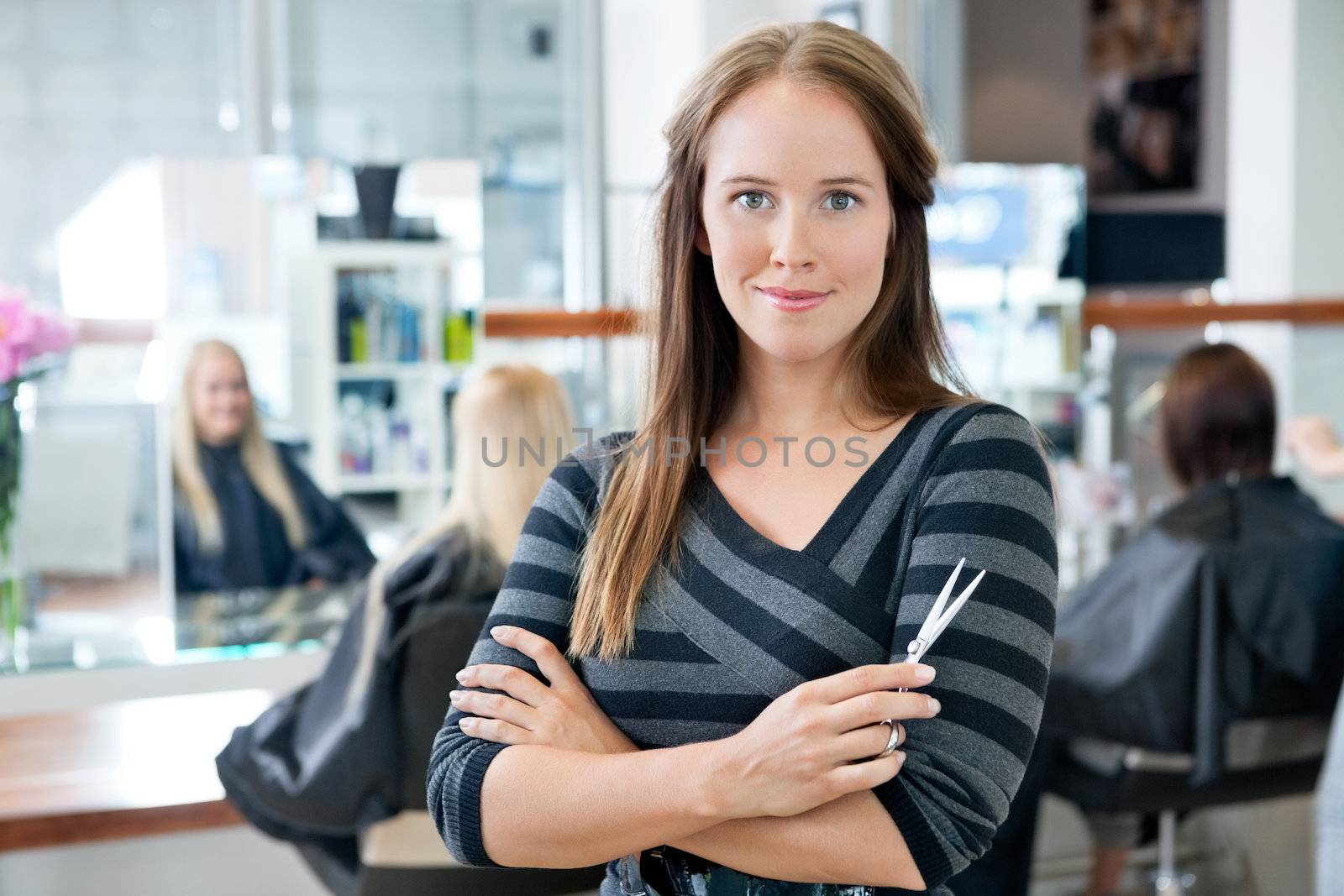 Portrait of a confident female hairdresser standing hands folded with people in background