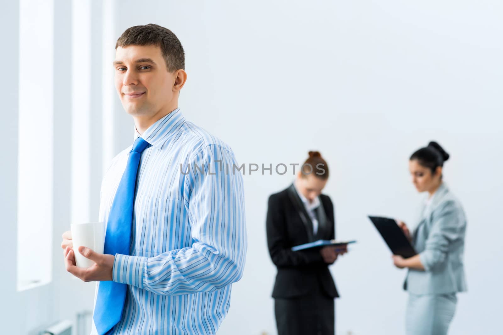 portrait of a successful businessman, holding a white mug, in the background talking colleagues