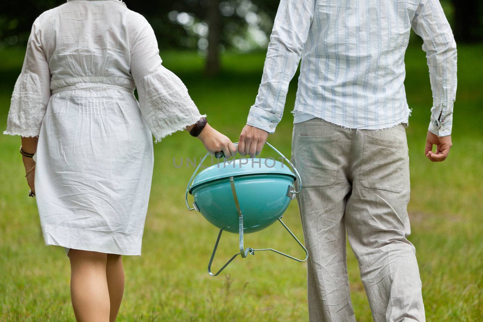 Back view of a couple in casual wear walking with a portable barbeque on an outdoor picnic