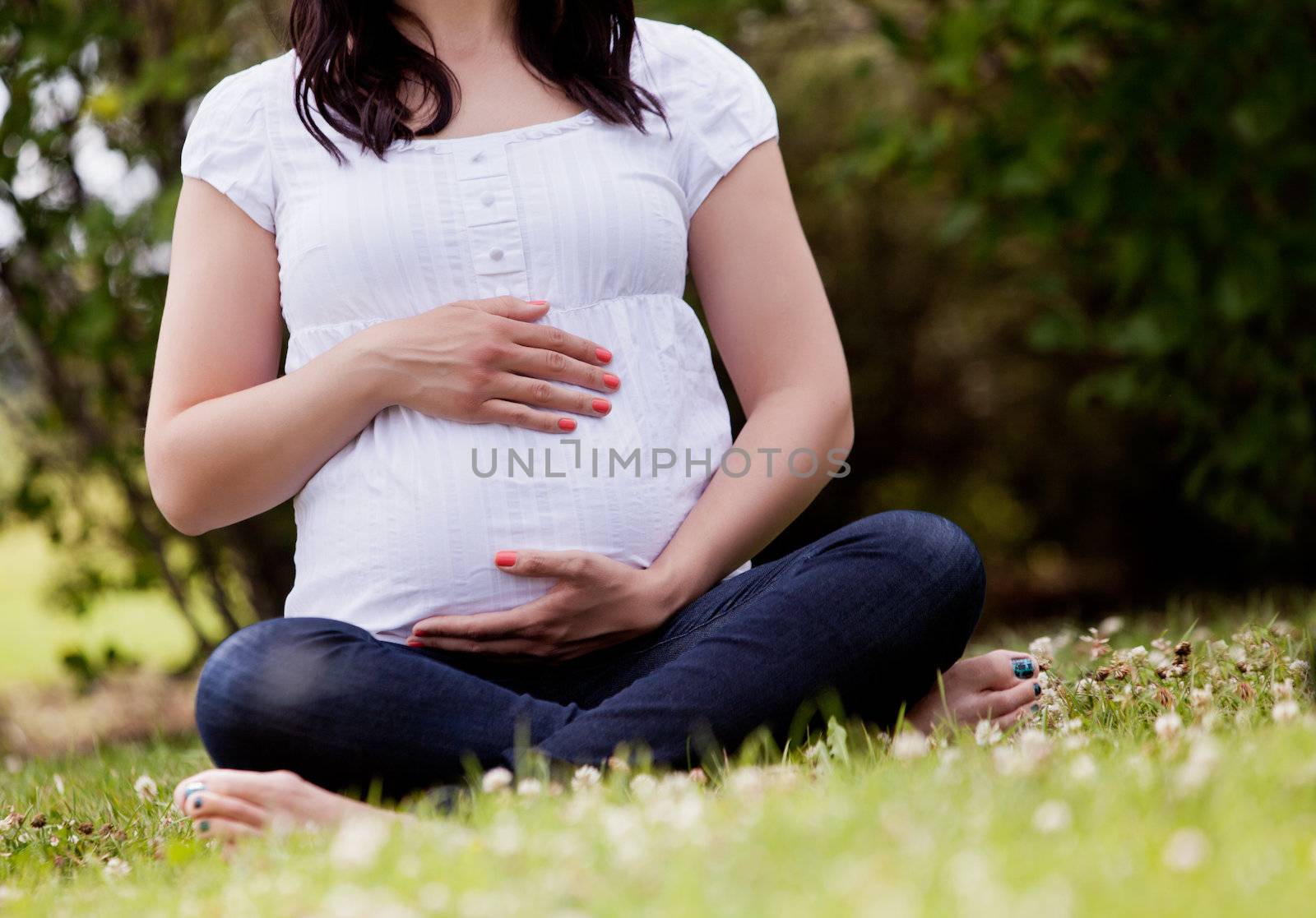 Faceless pregnant woman in third trimester sitting in park holding belly