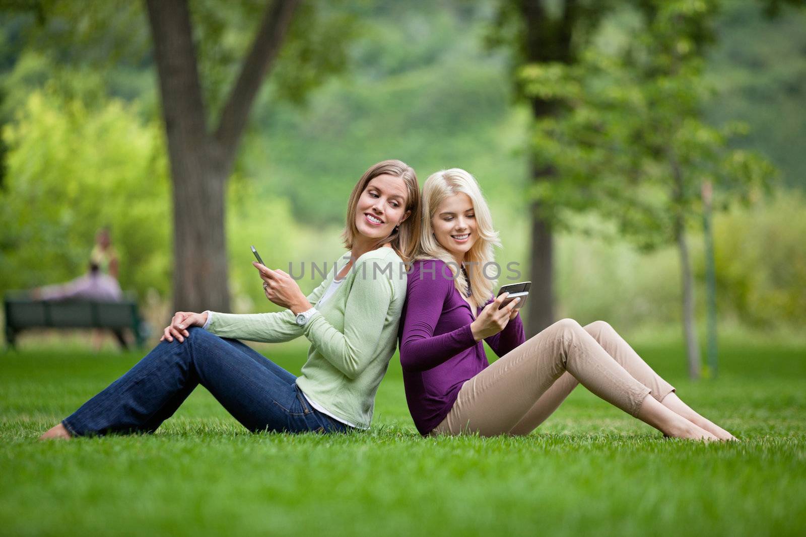 Side view of happy young female friends with cellphones sitting back to back in park