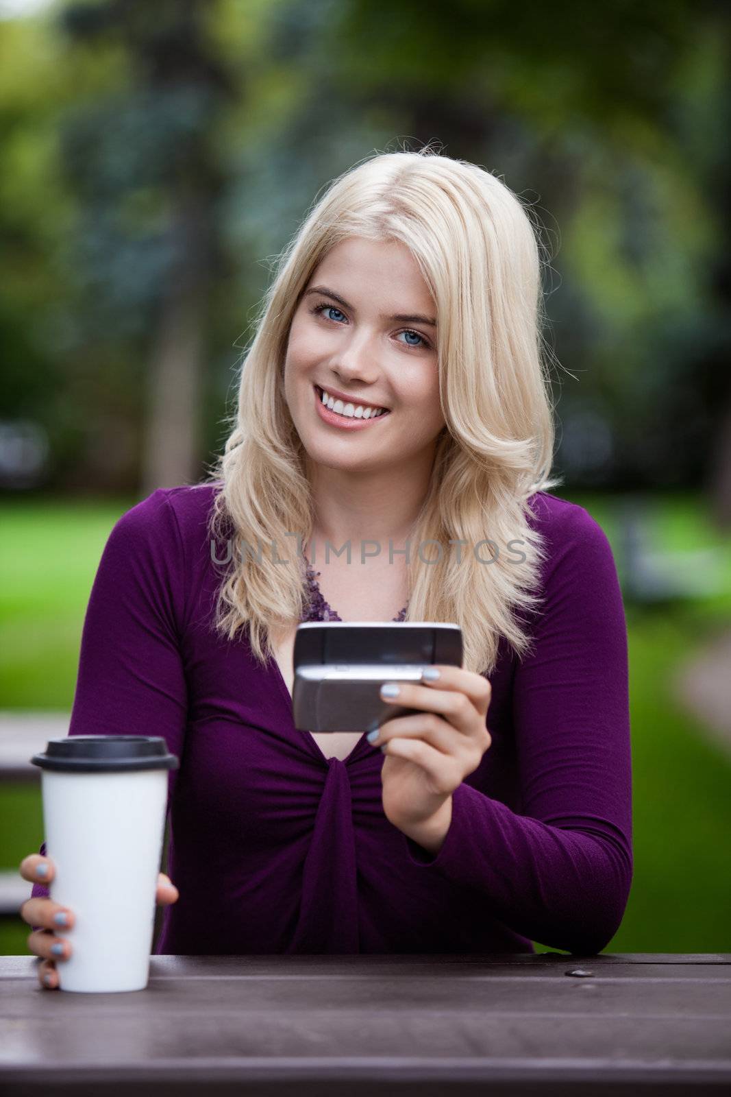 Portrait of a happy smiling college student sitting in park with cell phone and coffee