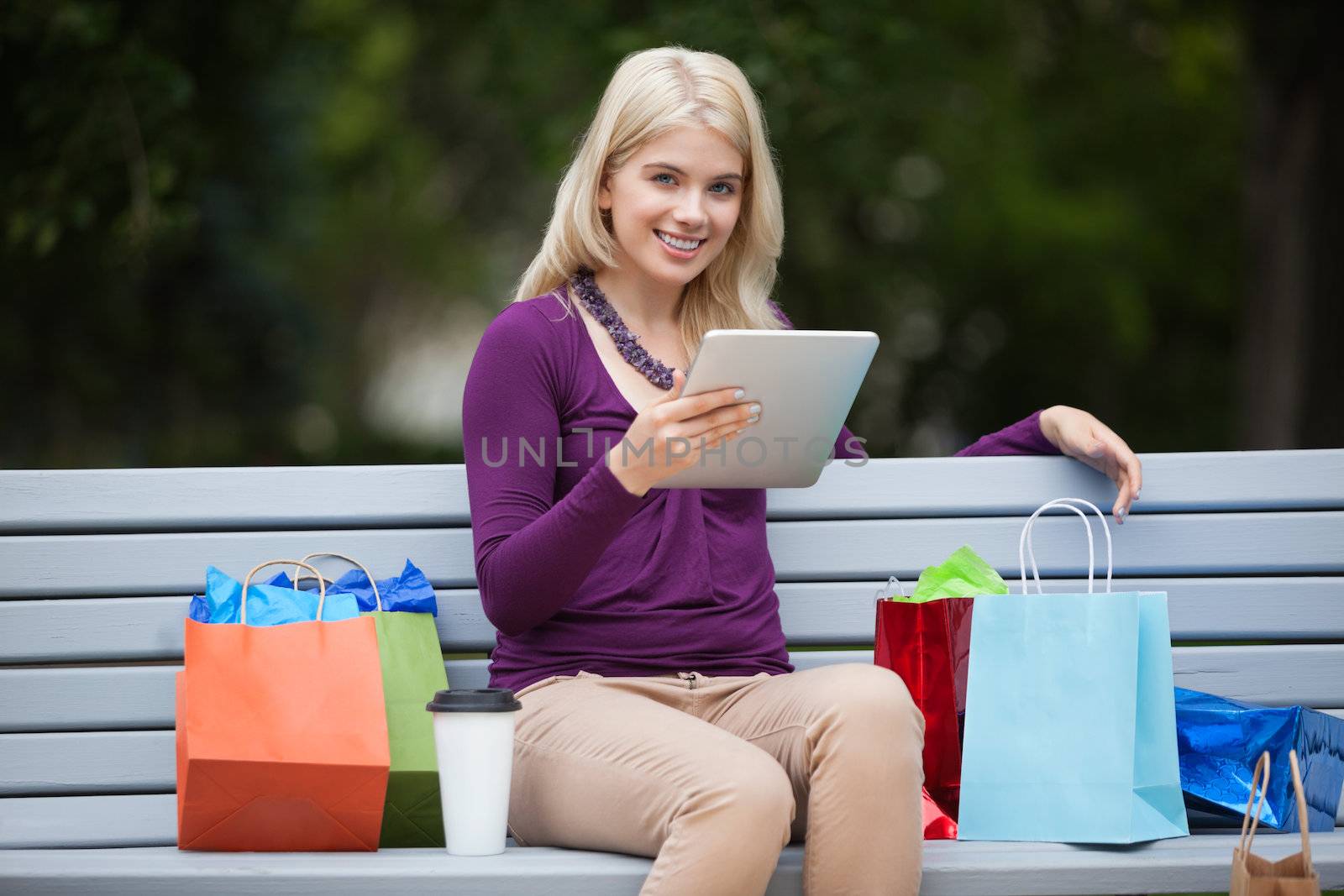 Portrait of pretty young woman with shopping bags using tablet PC on park bench