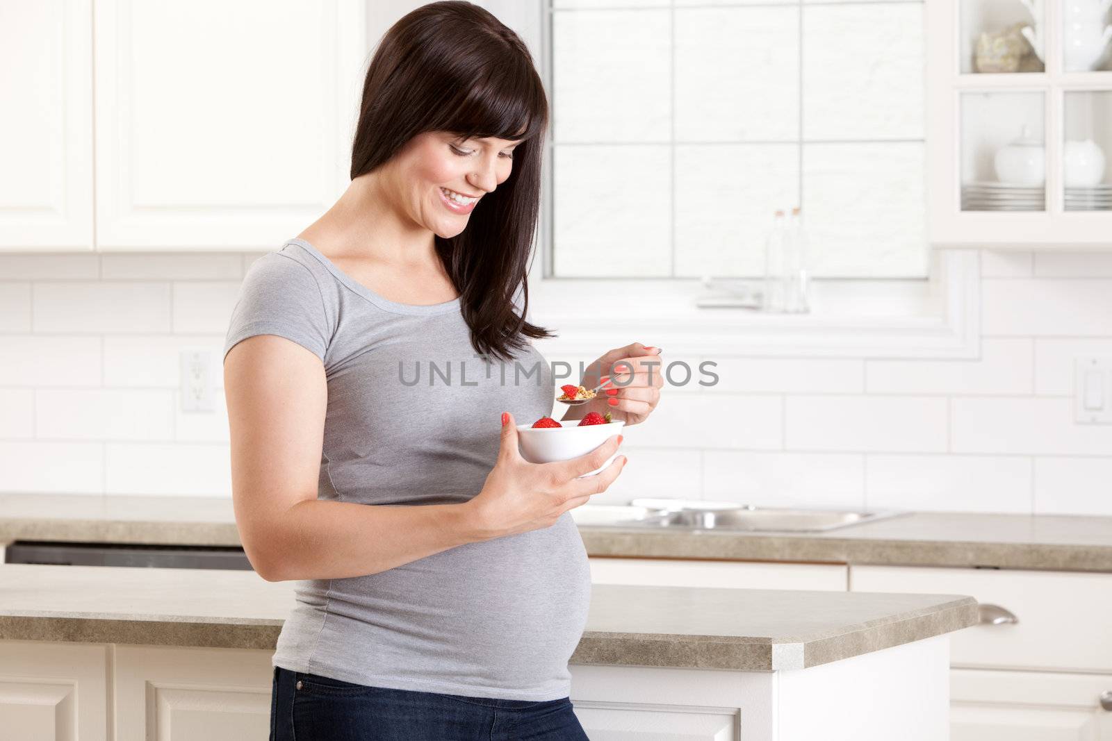 Happy pregnant woman in kitchen eating a healthy snack of strawberries and granola
