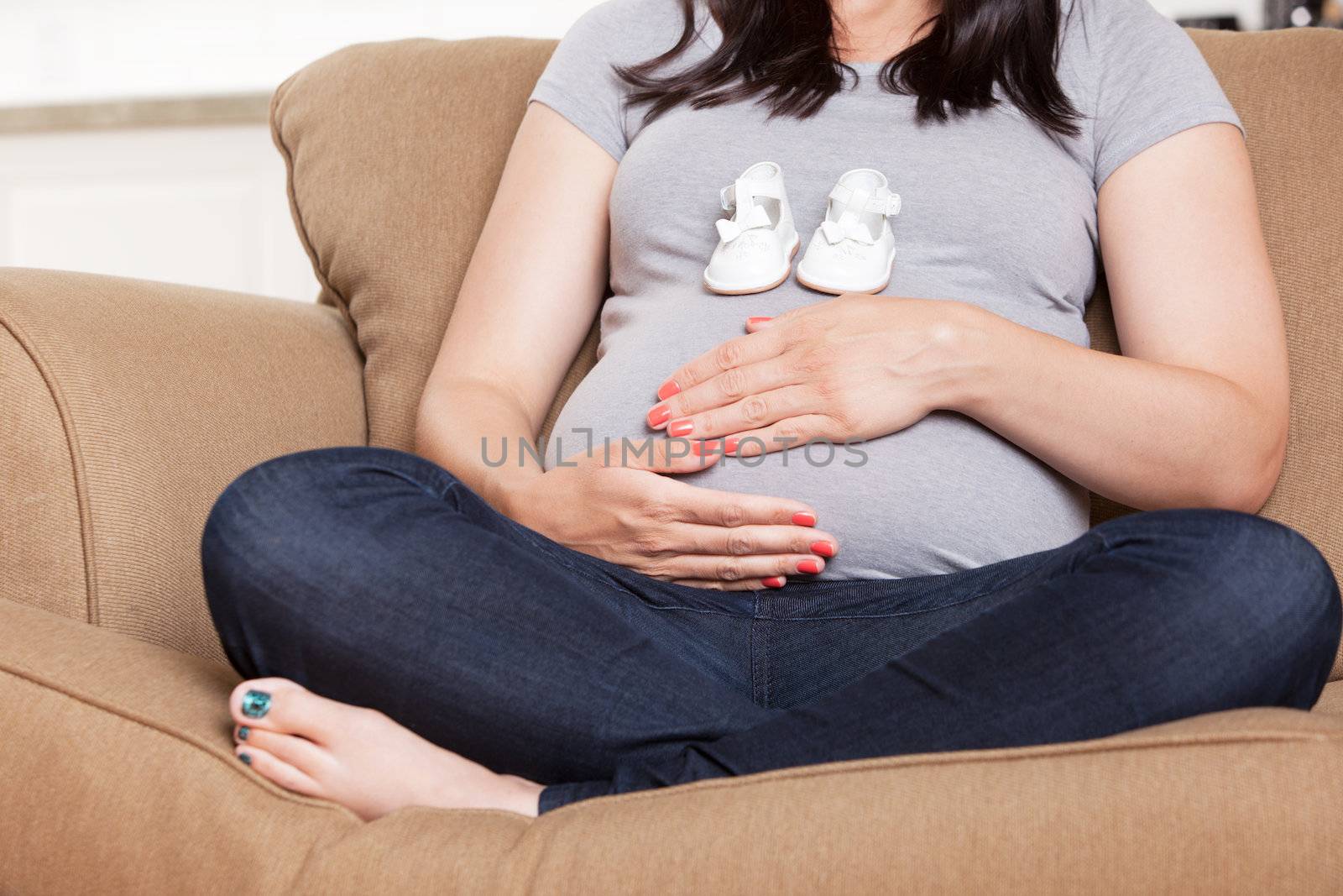 Faceless young pregnant woman on sofa holding baby shoes on belly
