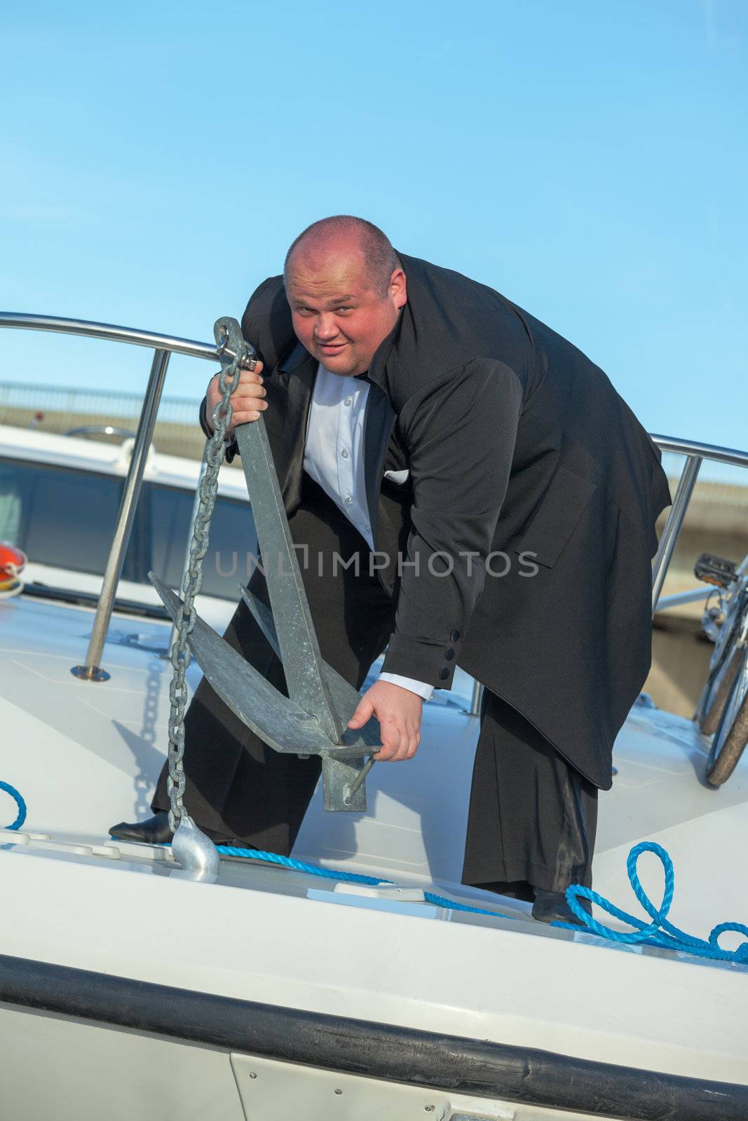 Overweight man standing on the deck of a luxury pleasure boat wearing a suit and lifting an anchor, conceptual of wealth