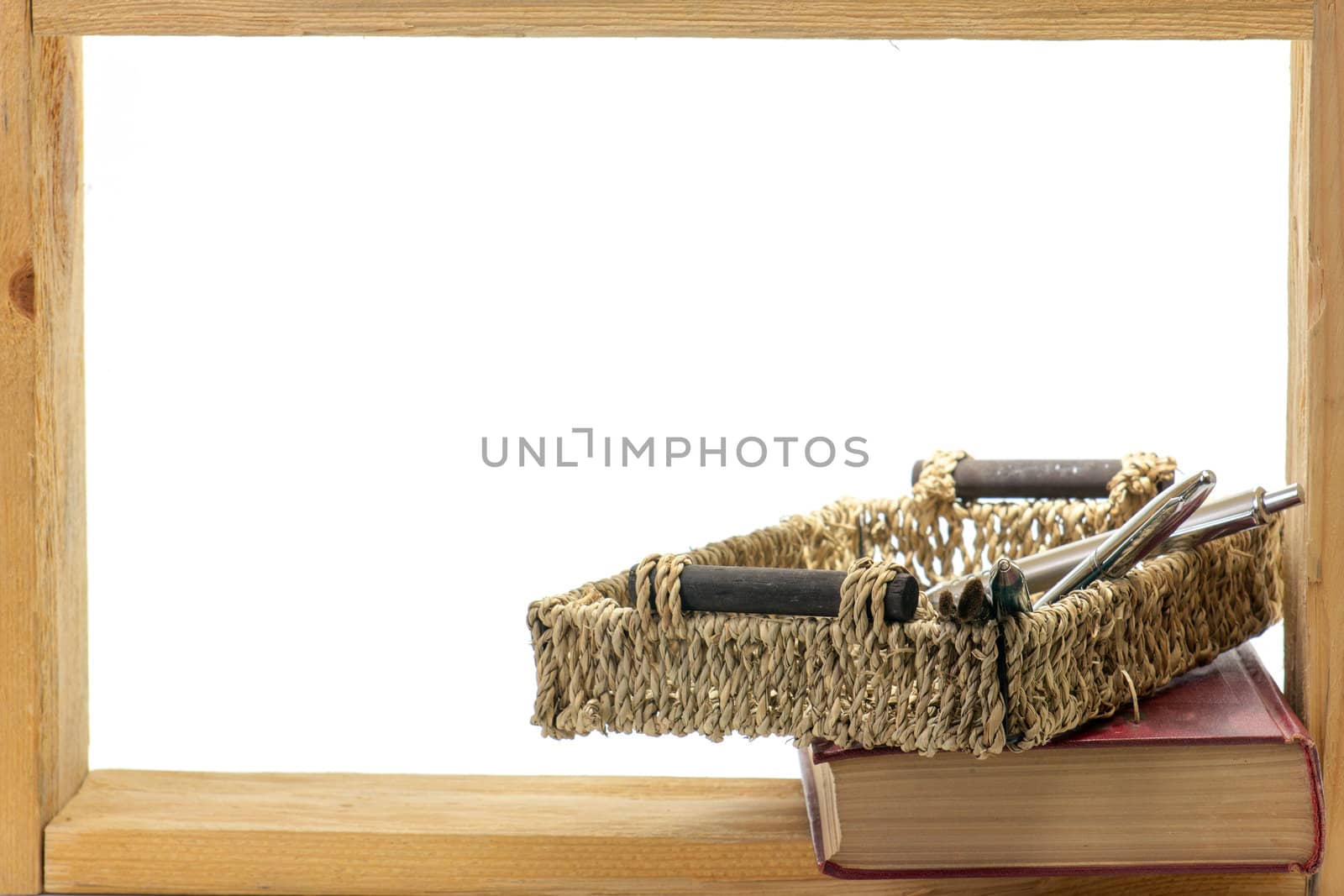 Book and wicker basket with a ball-point pens and brushes in a wooden frame on white background