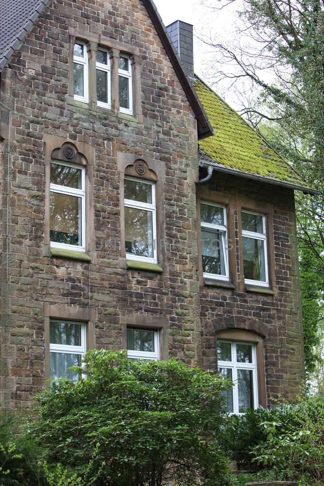 View of a multistorey old stone house with a green mossy tiled roof partly hidden behind green vegetation