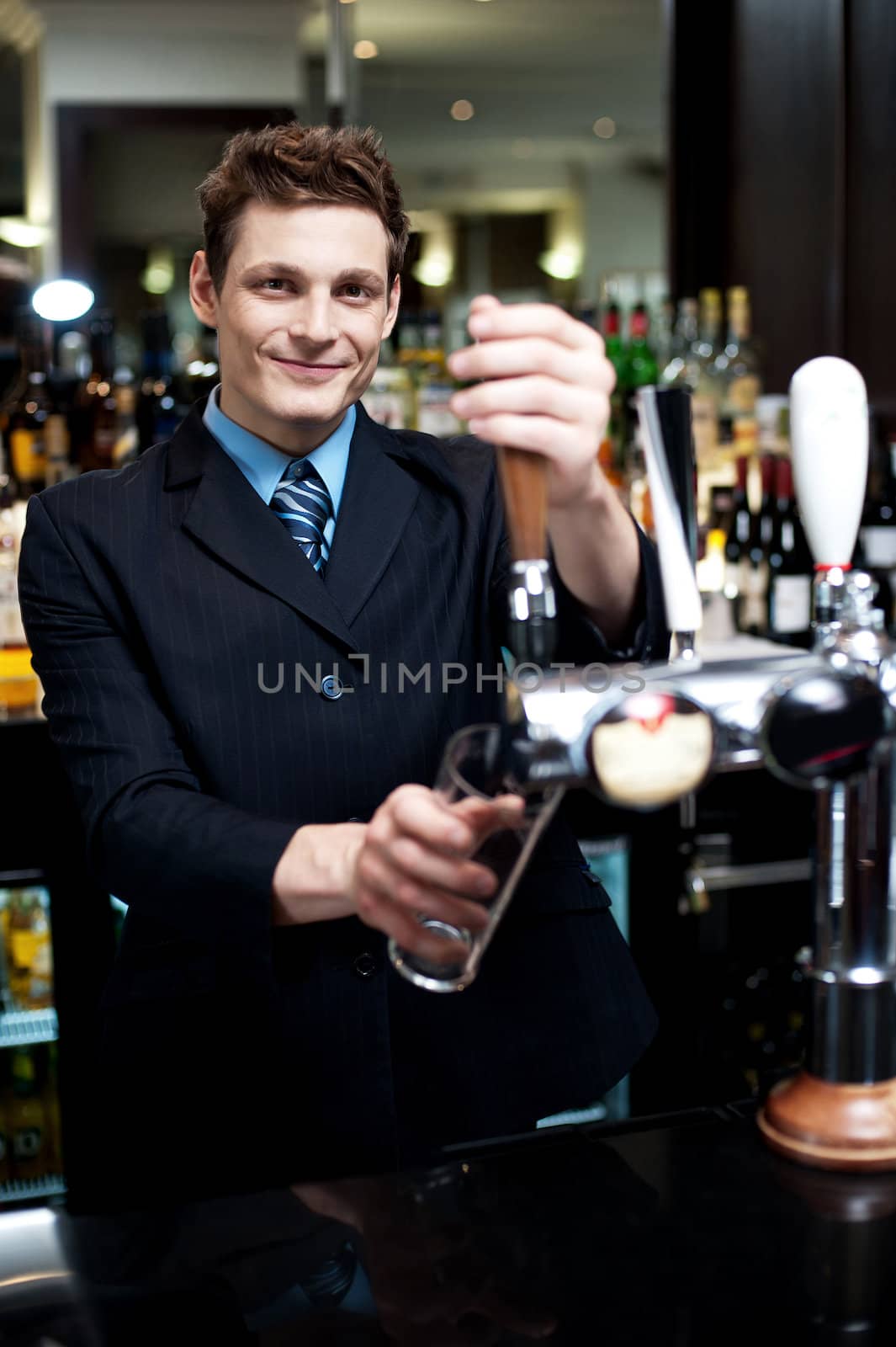 Handsome mixologist putting ice into tall glass. Bar decorated with bottles in the background