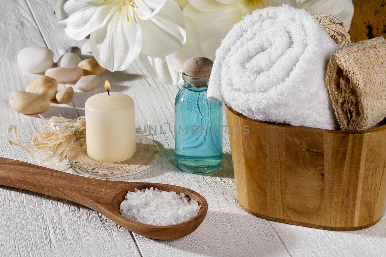 A set of spa products with spa salt on a spoon, towel, oil and a lighted candle on a wooden table