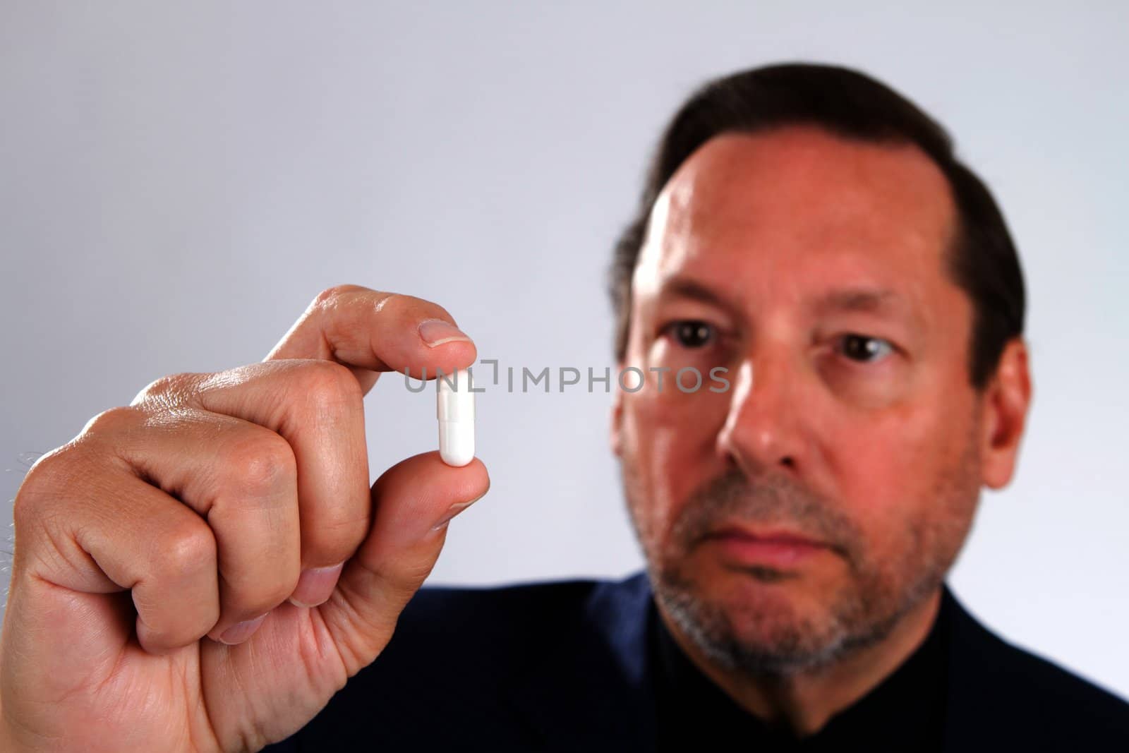 Senior adult man holds medicine capsule in his hand with him in the background out of focus and focus is on the capsule.