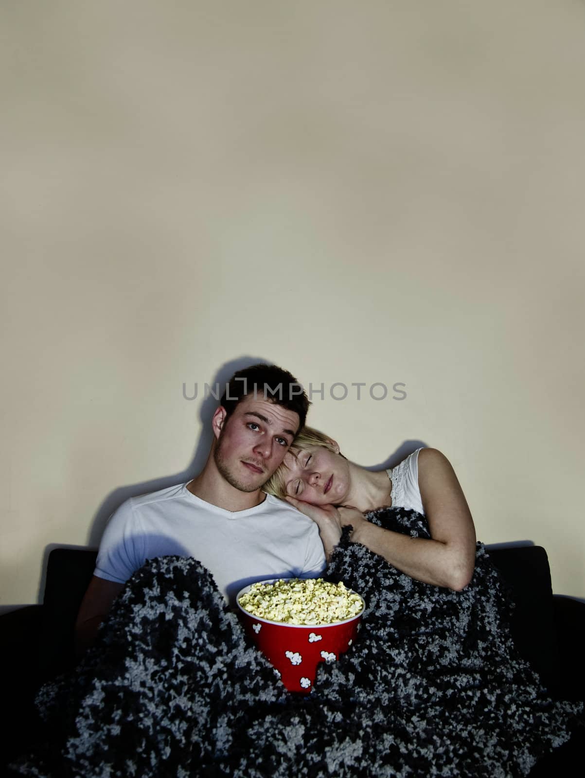 Young couple watching television and eating a huge bowl of fresh buttered popcorn comfortably wrapped in a blanket. The movie might be a bit boring since both of then are sleeping... in the popcorn !!!