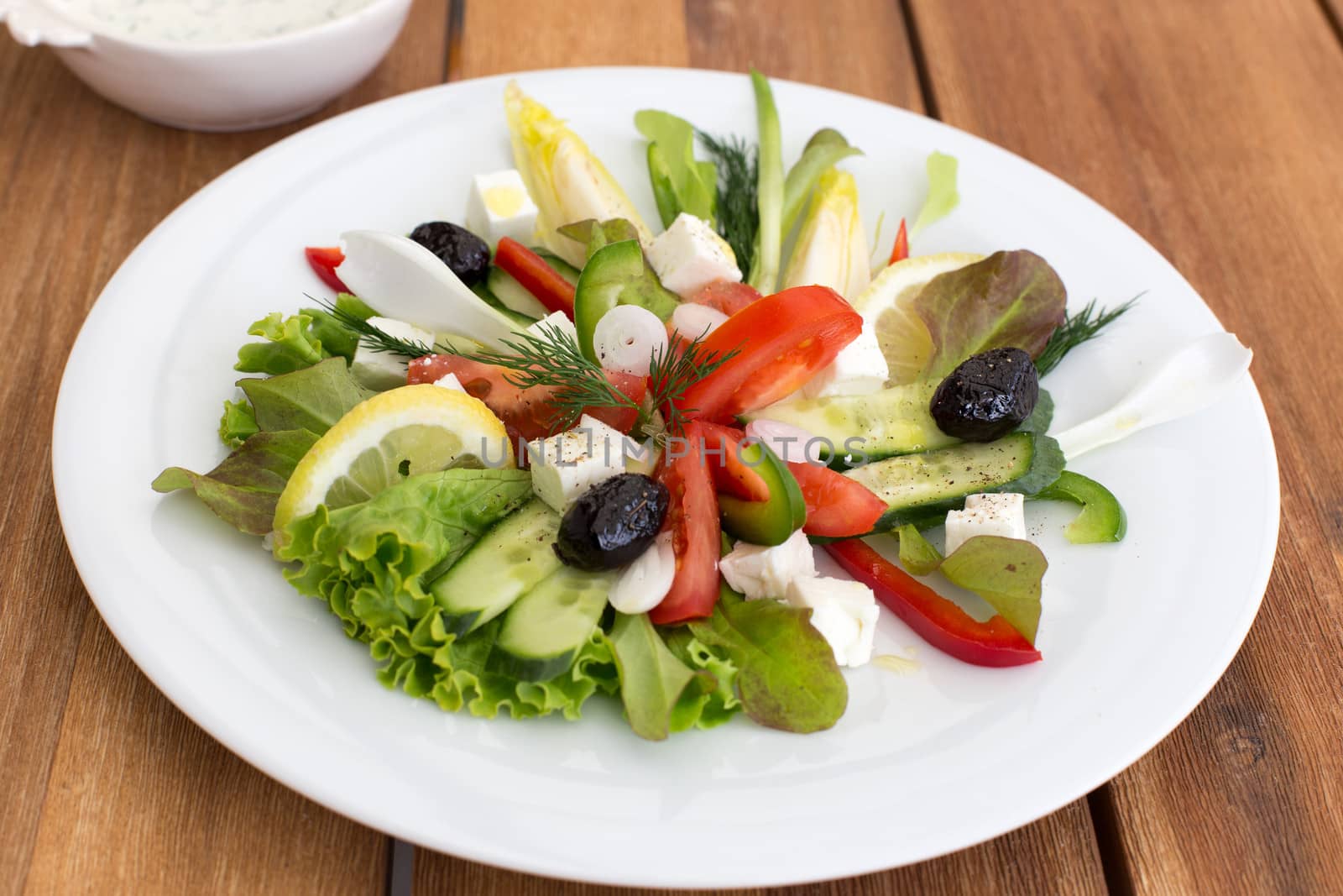 Greek salad with feta, tomatoes, cucumber, peppers and black olives. With bowl of sauce on a wooden table.