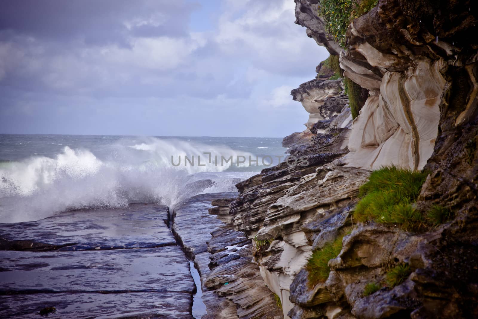Waves breaking on a wide rocky shelf at the foot of a cliff under a cloudy summer sky