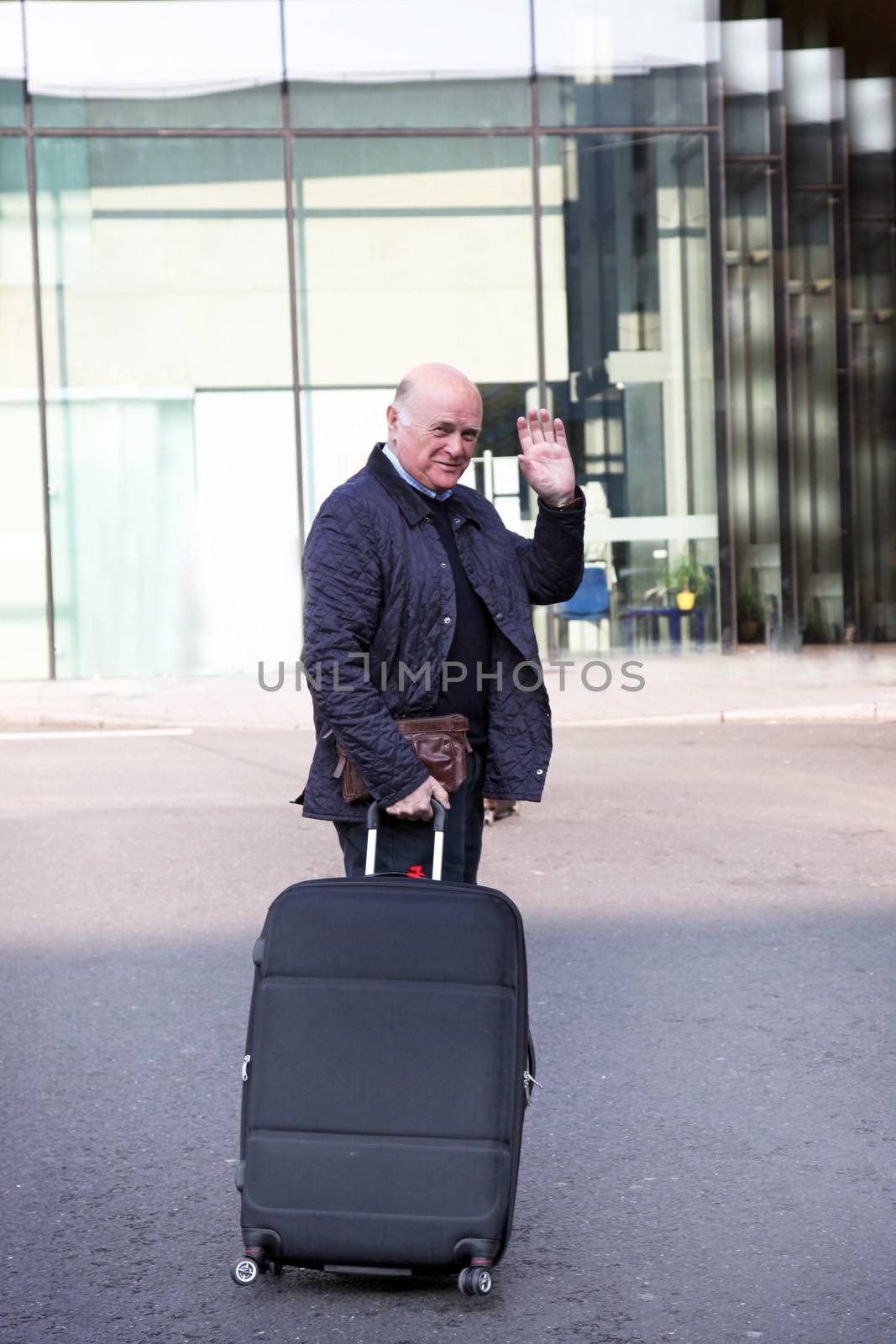 Elderly man waving goodbye as he makes his way to the departure terminal with his suitcase at the end of his holiday