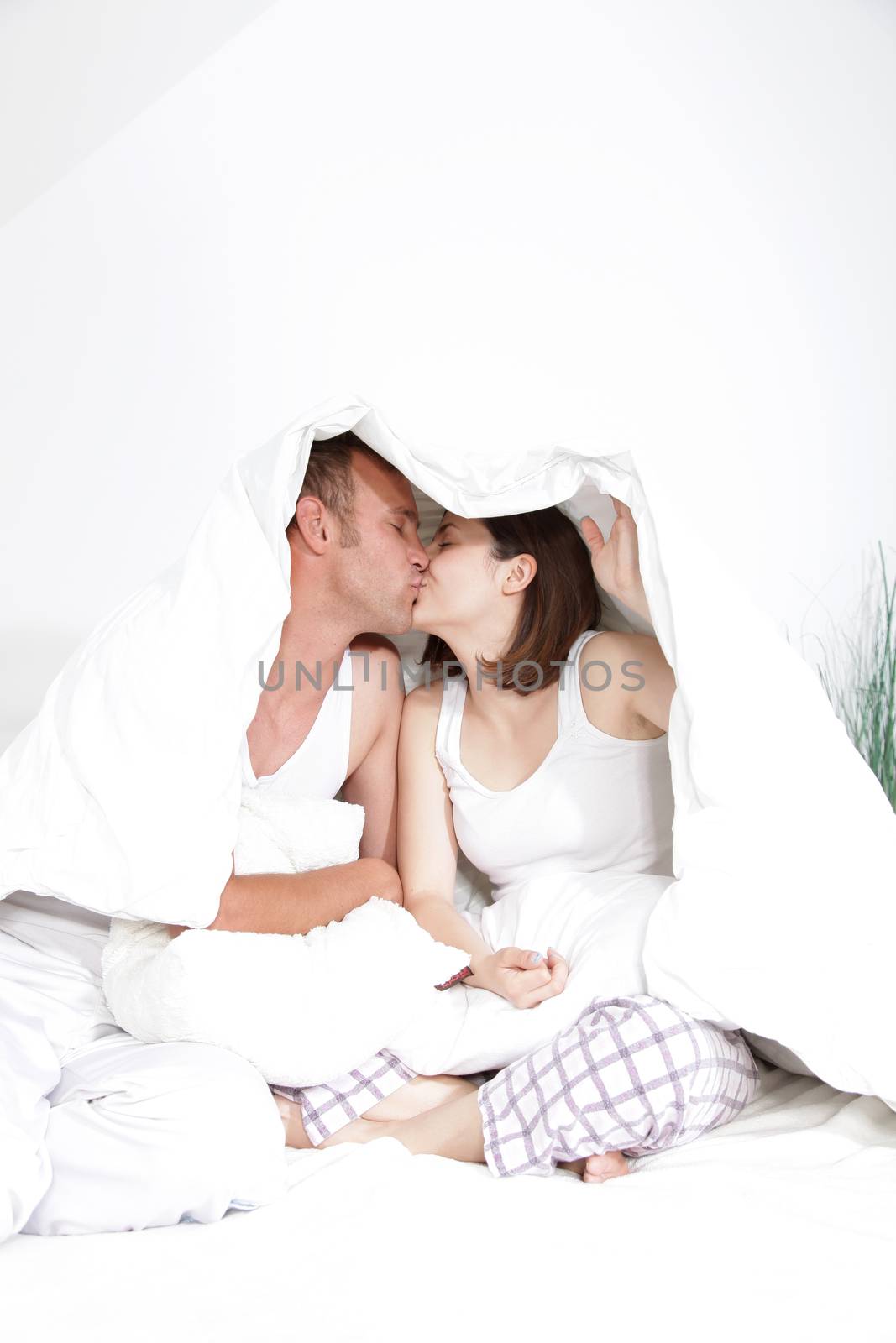 Romantic young couple sitting on the bed kissing under the bedclothes with the duvet over their heads